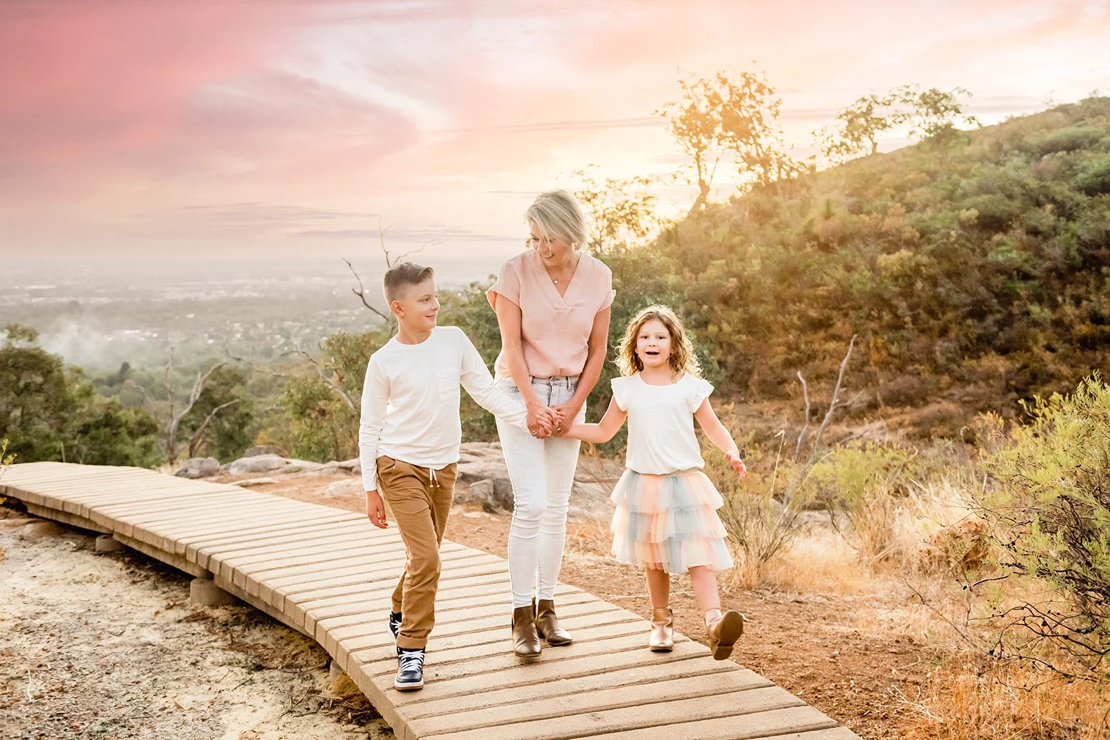 A woman with two children walking on a wooden trail during sunset, holding hands and smiling, with a scenic view of a valley and hills in the background.