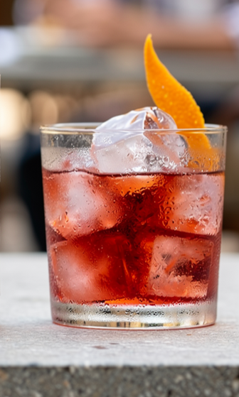 Un bicchiere di Bitter and Soda preparato con il cocktail BOTANICA Bitter Zero con cubetti di ghiaccio, guarnito con una fetta di scorza d'arancia, su una superficie bianca.