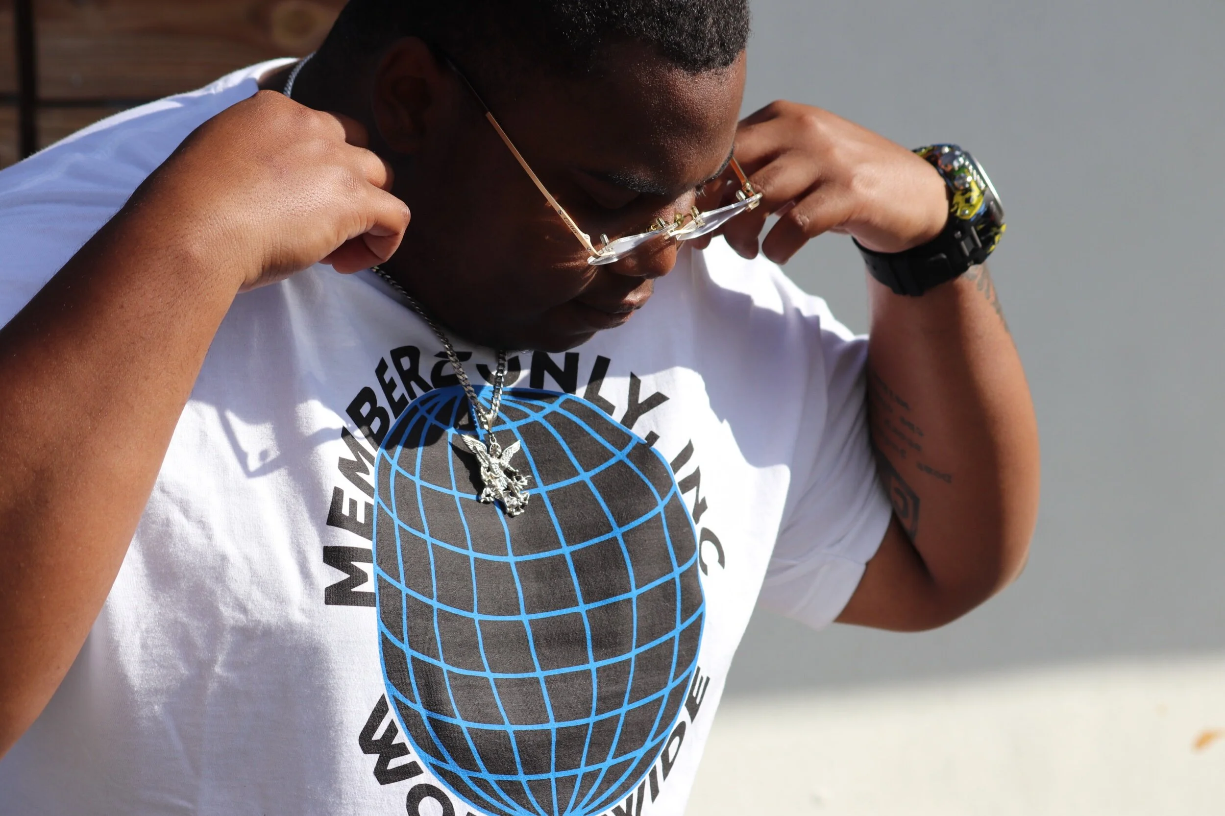 A person wearing a white t-shirt adjusting their glasses, with a necklace featuring a winged animal pendant. The t-shirt has a globe graphic with the words 'MEMBER ONLY' and 'WORLDWIDE' around it.