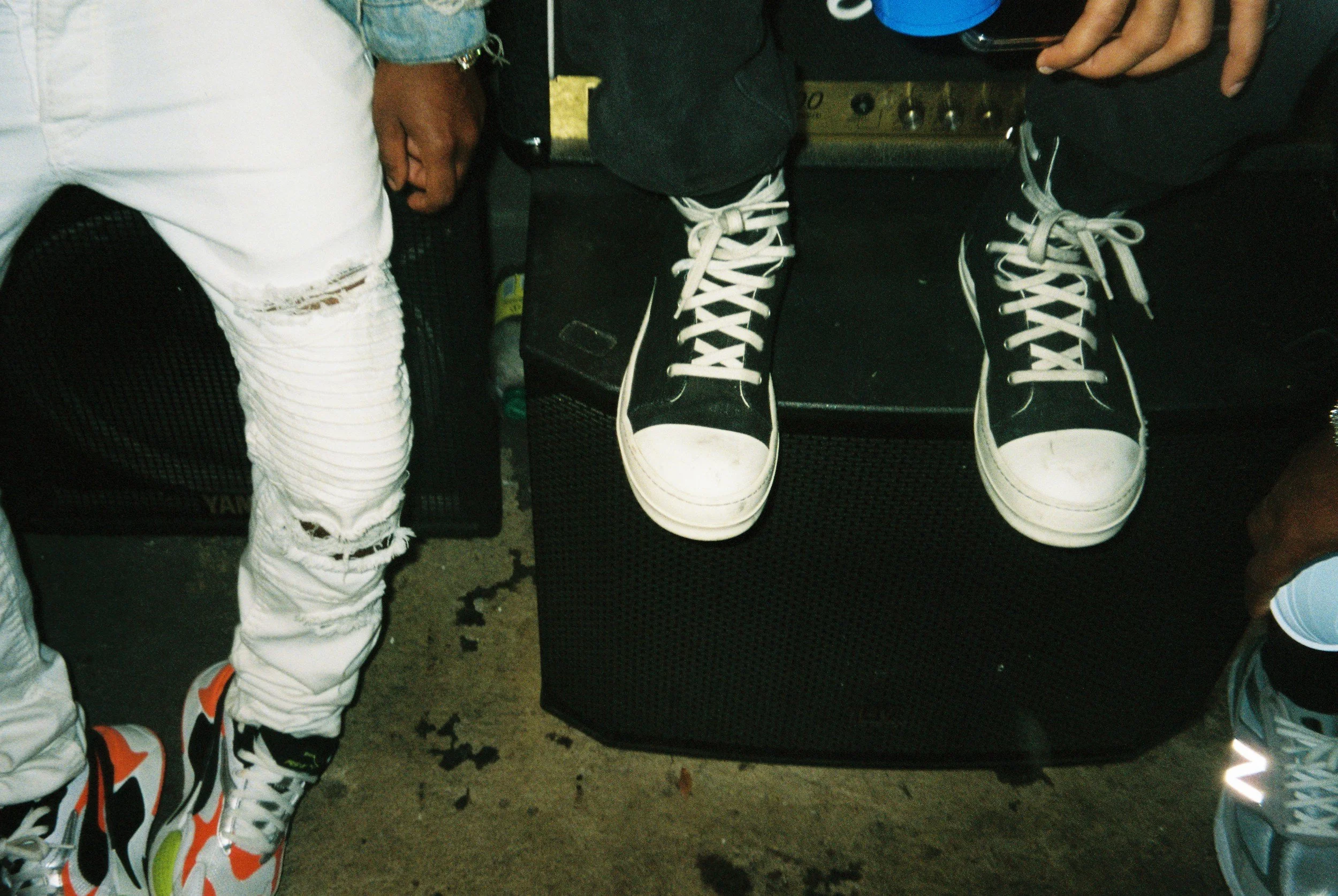 Close-up of two black and white sneakers on a black speaker, with two people sitting nearby. One person is wearing white ripped jeans and colorful sneakers, and the other wearing dark pants and blue sneakers.