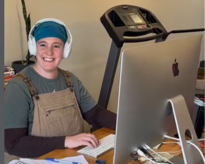 Woman with earphones on and blue scarf in overalls sitting at an apple computer smiling because she loves SEO and now she gets to talk about it.
