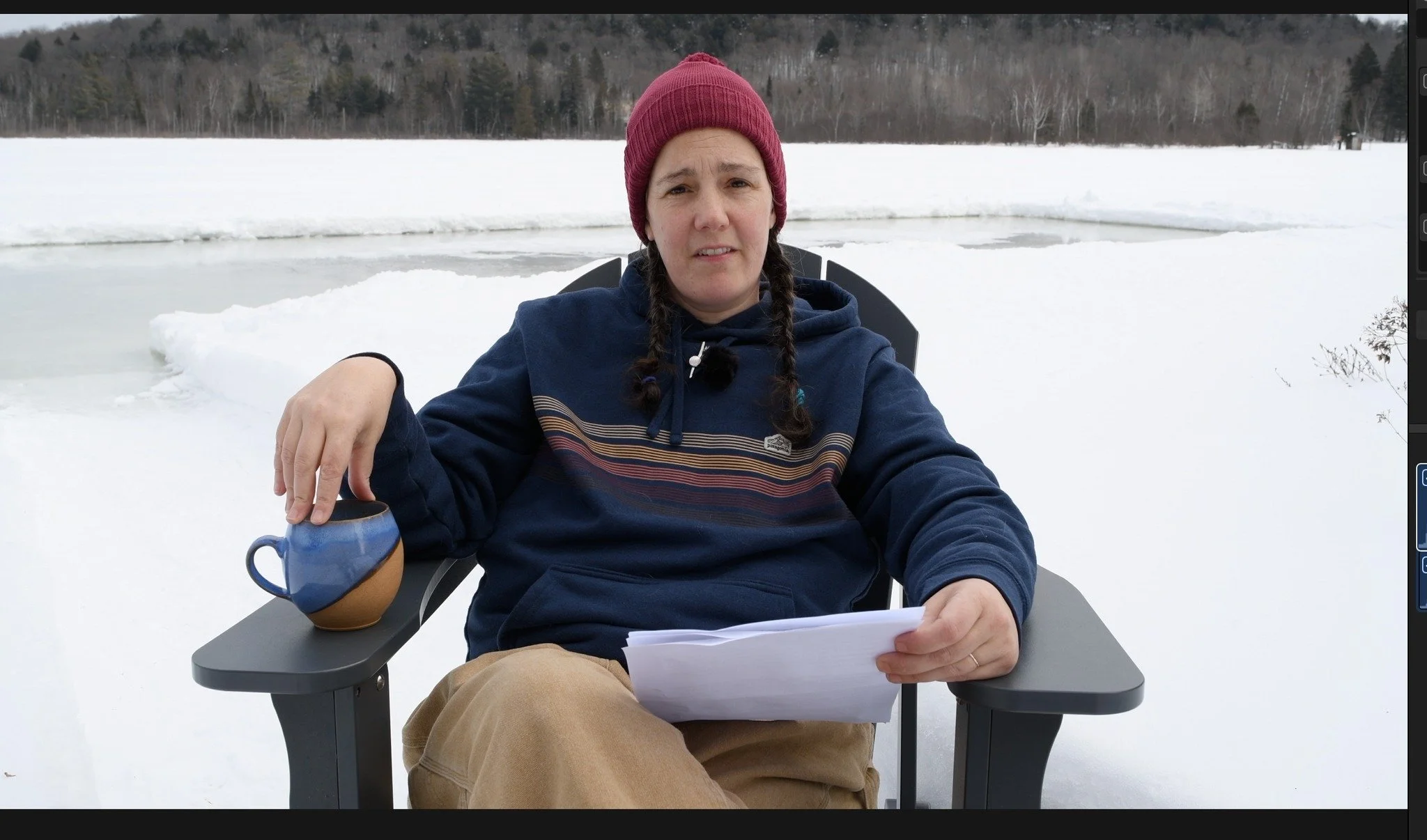 Woman in red tuque, blue sweatshirt, sitting on a chair outside in the winter. She has a blue pottery coffee cup and paper in her hands.
