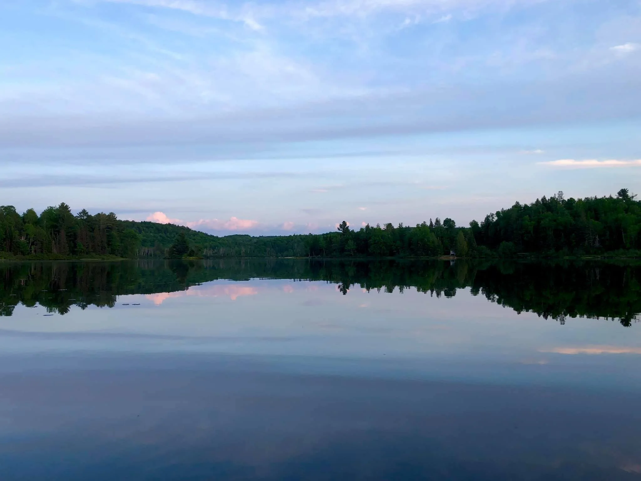 Blue and pink clouds at sunset over a lake. There are trees and a beautiful reflection of the sky on the lake.