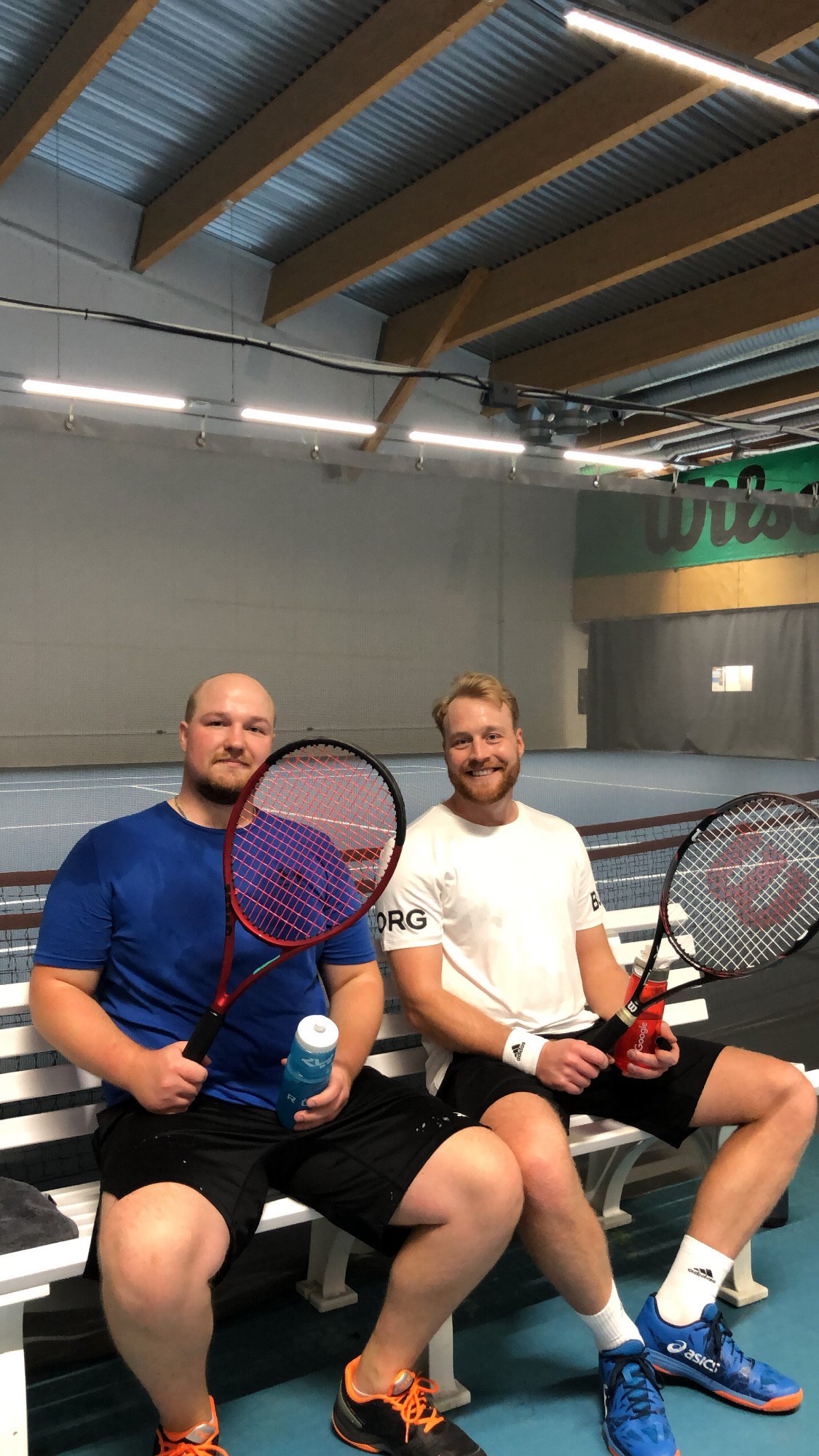 Kaksi miestä istuu penkillä tennistunnin jälkeen, hymyilee kameralle ja pitää mailoja ylhäällä.