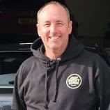 A man smiling outdoors at night, wearing a black hoodie.