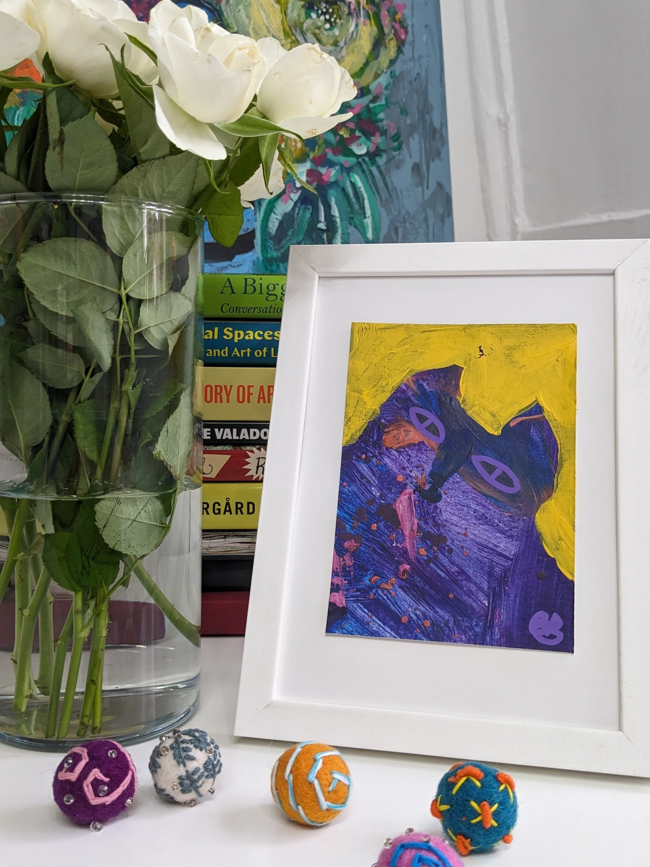 A white-framed abstract painting with purple, yellow, pink, and orange colors along with purple circle shapes. A vase with white roses is on the left side, and several books are behind the vase. Colorful felted balls with embroidery are on the bottom in front of the painting.