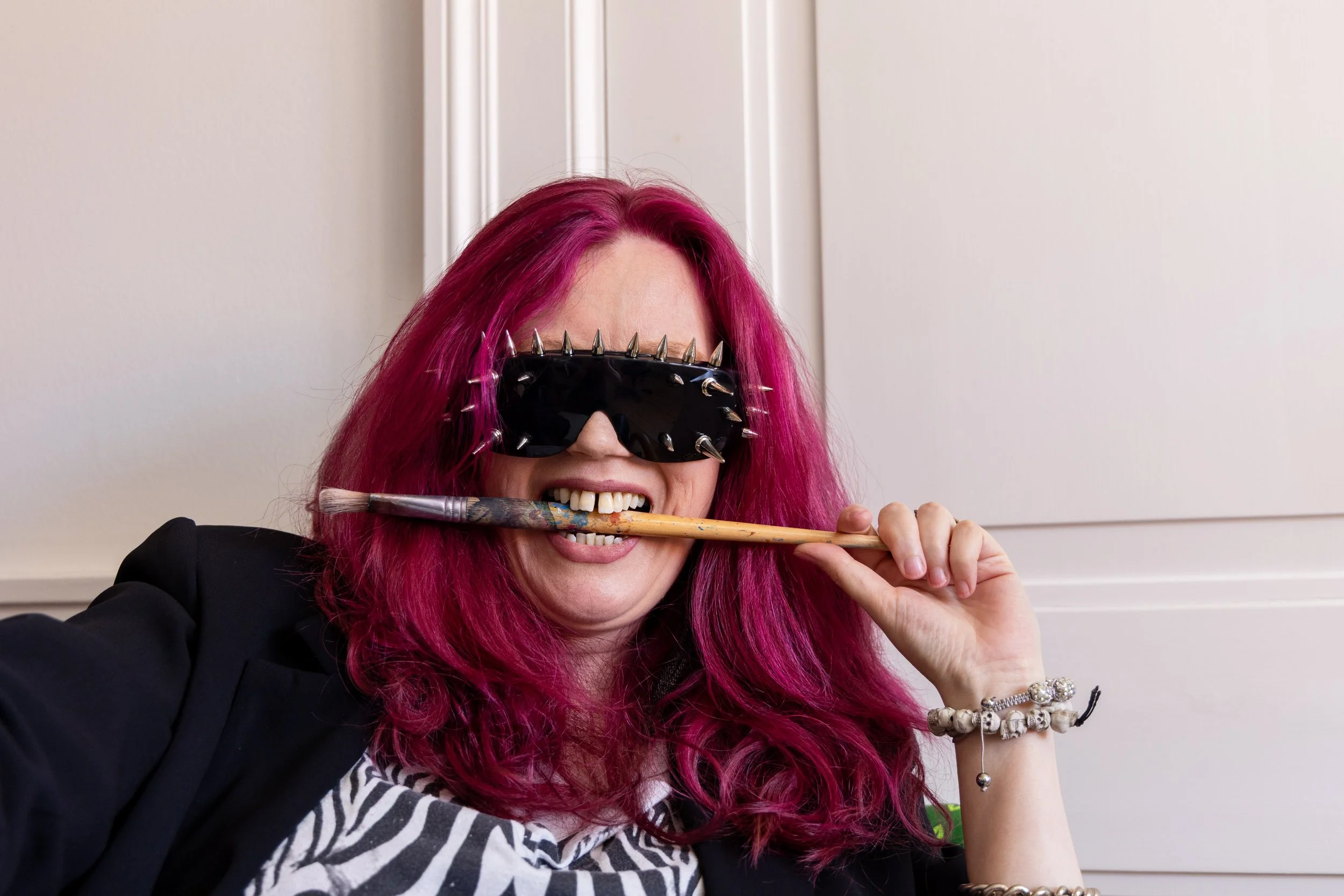 A woman with bright pink hair wearing spiked black sunglasses, holding a paintbrush in her mouth, and smiling.