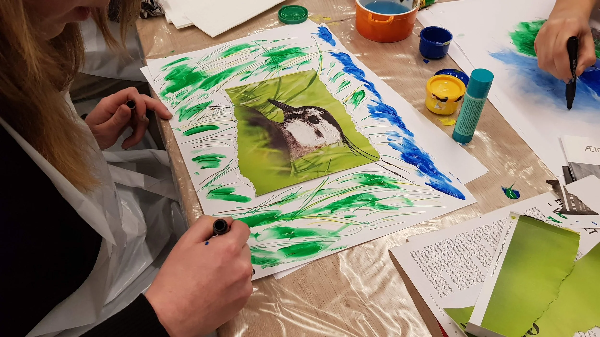 People creating an artwork featuring a bird photograph surrounded by painted green grass and blue sky on paper.