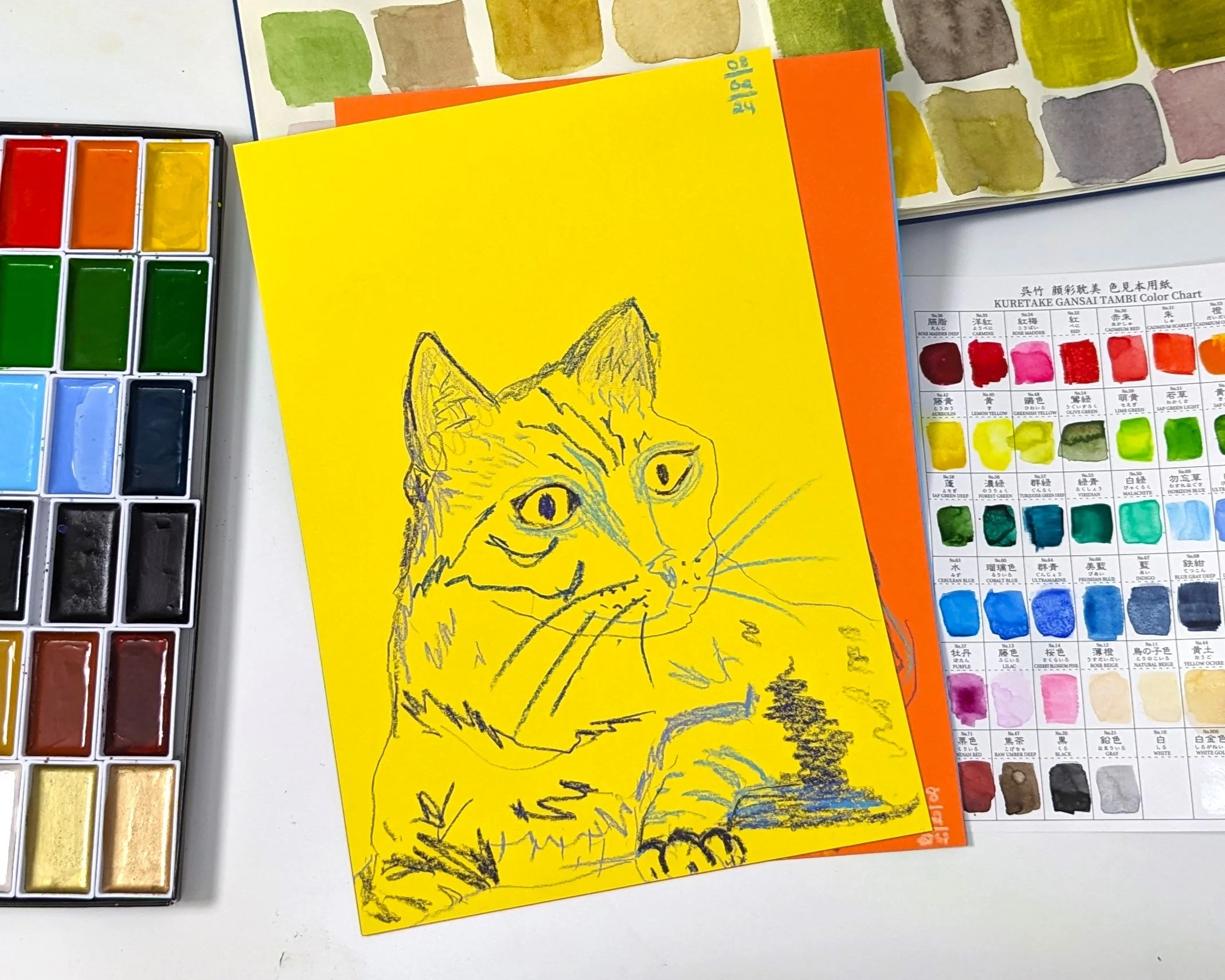 A yellow piece of paper with a sketched drawing of a cat on it. The drawing is done with black and blue ink, showing detailed fur and facial features. Surrounding objects include watercolor paints, color charts, and part of a watercolor paper with color swatches.