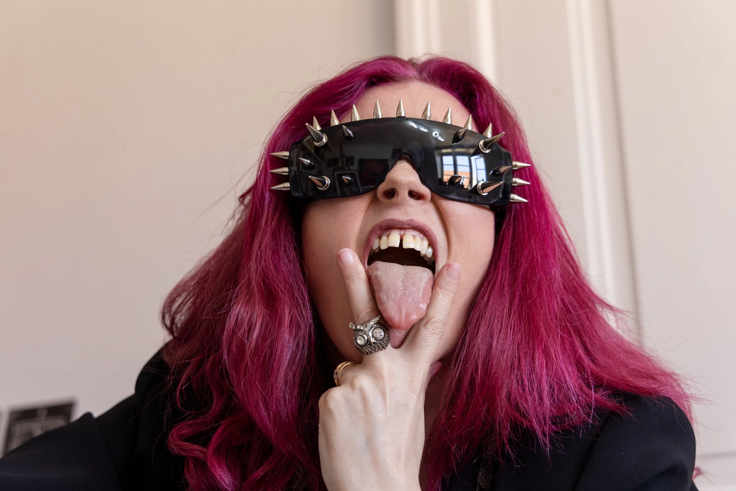 Person with bright pink hair wearing black sunglasses decorated with silver spikes, sticking out tongue, and holding chin with hand.