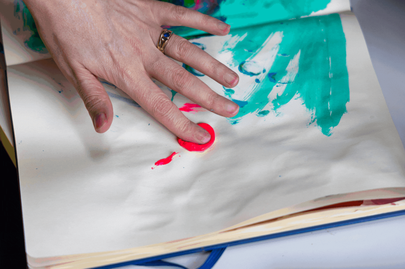 A person's hand with a ring is touching a page in an art book, which has colorful abstract paintings with pink and blue paint.
