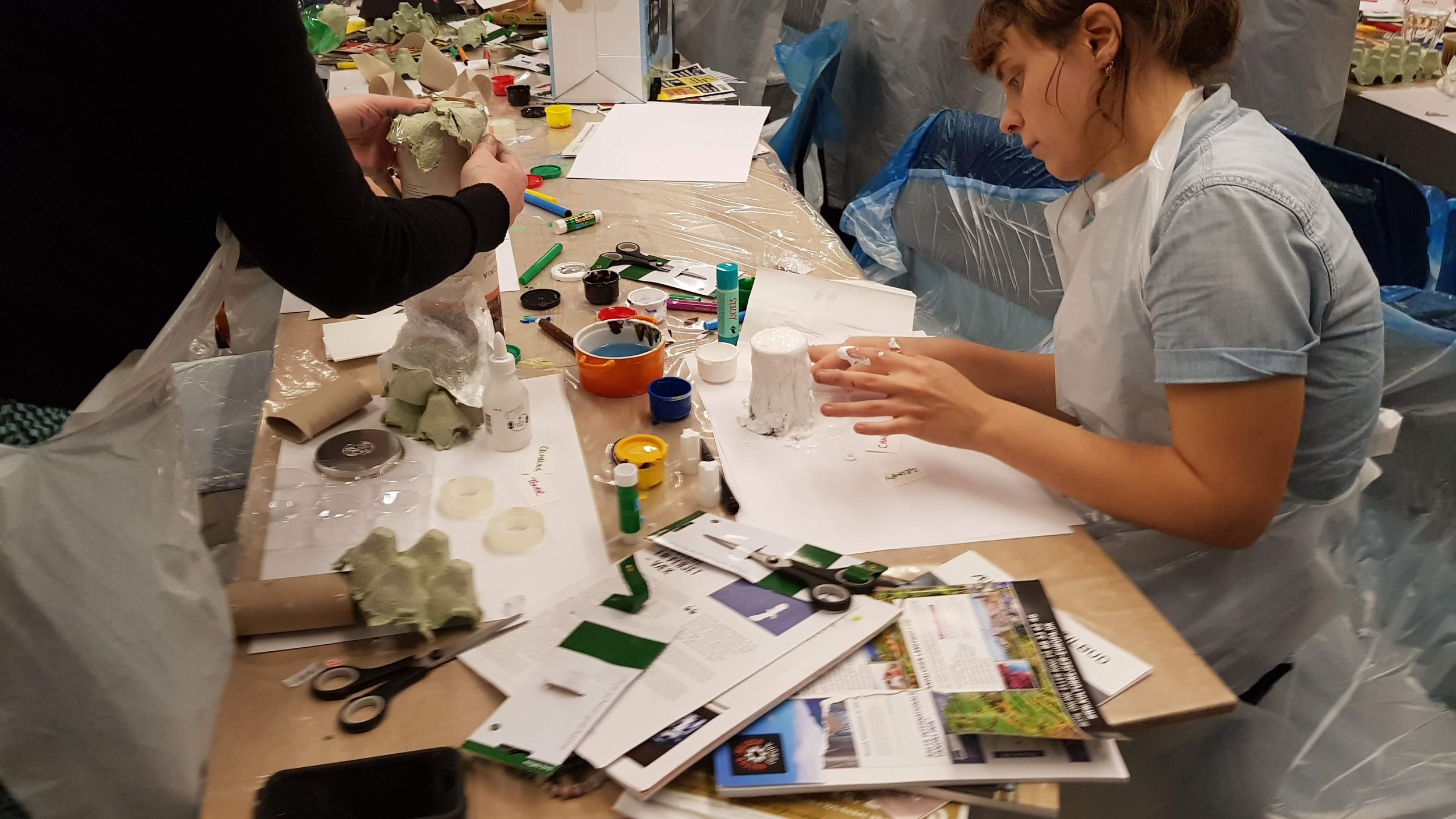 Two women working on a craft project at a cluttered table with papers, scissors, paints, and tools in a workshop or classroom setting.