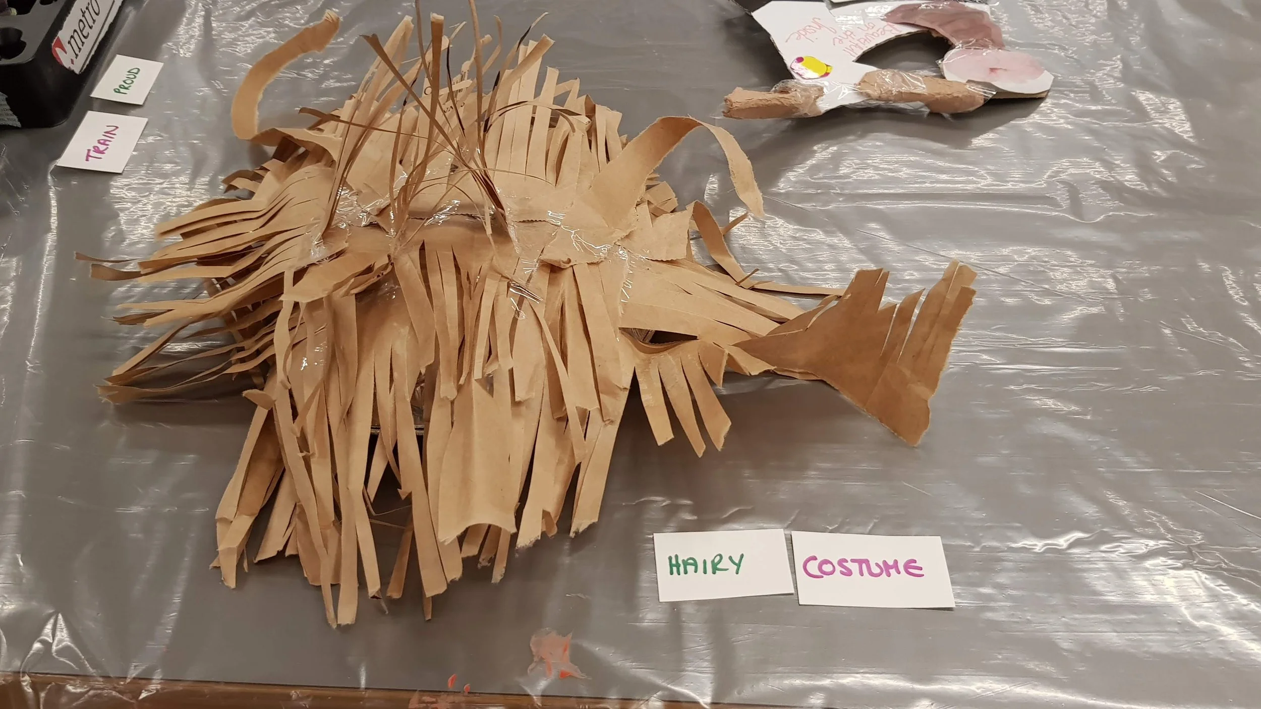 A craft project of a lion made from tan paper, with leaves for a mane, on a table with labels reading 'HAIRY' and 'COSTUME'.