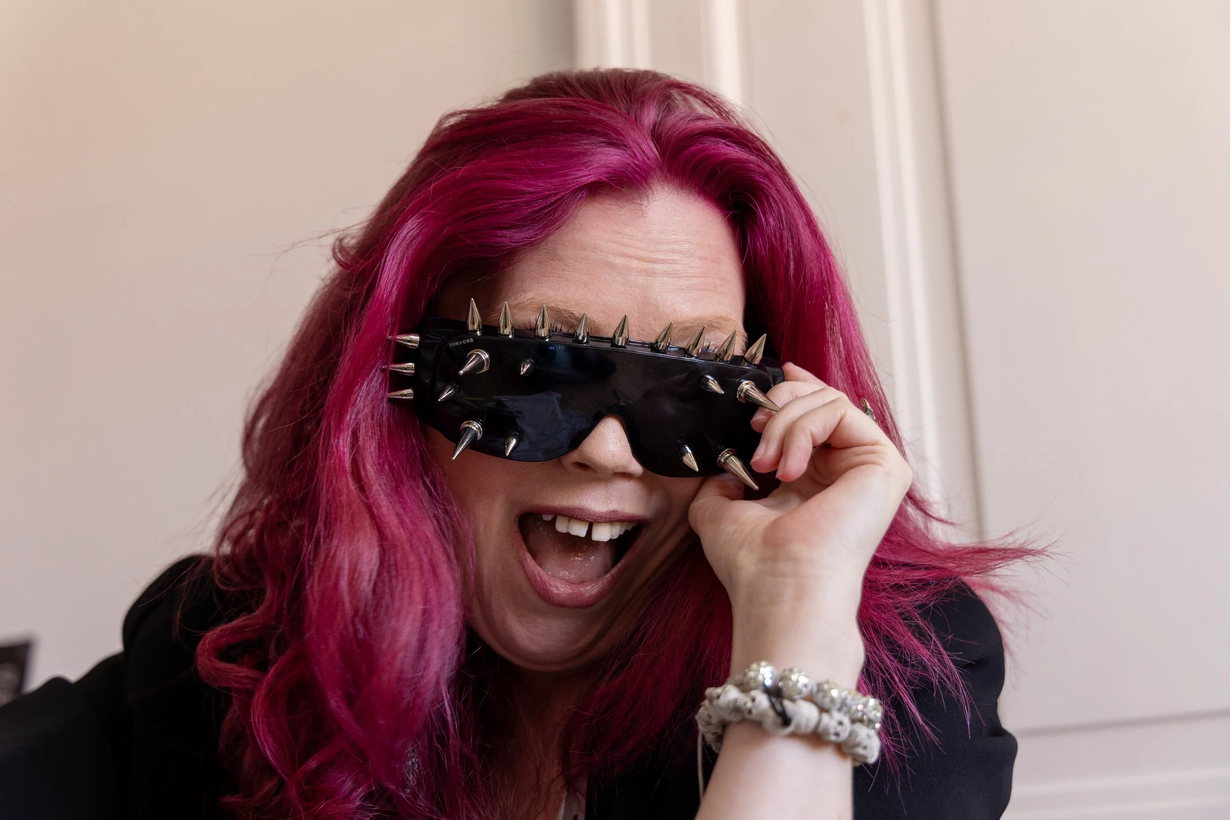 Woman with vibrant pink hair wearing spiked black goggles, smiling and laughing.