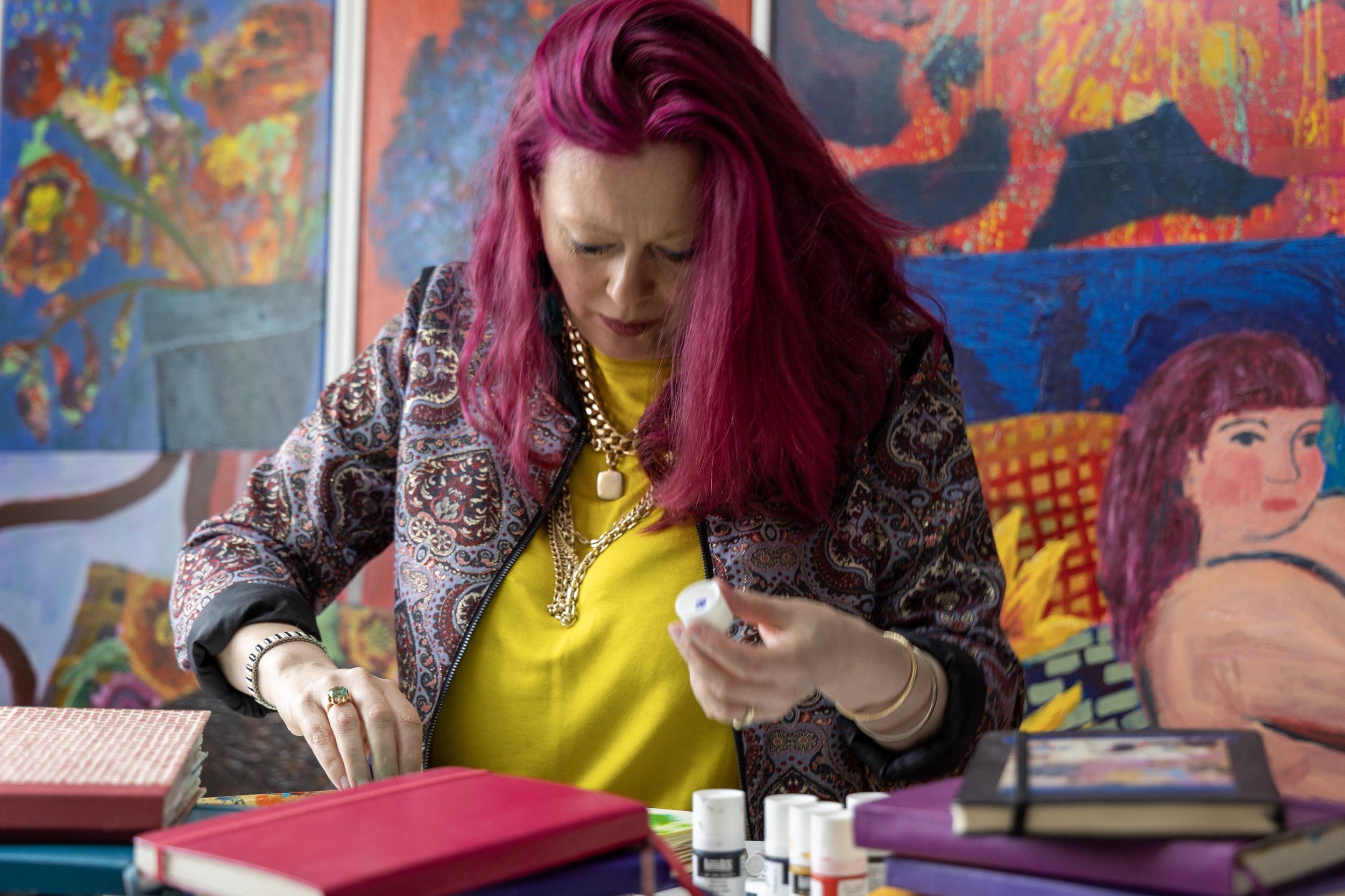 A woman with pink hair, wearing a patterned jacket and gold jewelry, is sitting at a table with books and art supplies, looking down at an item in her hand, with colorful paintings in the background.