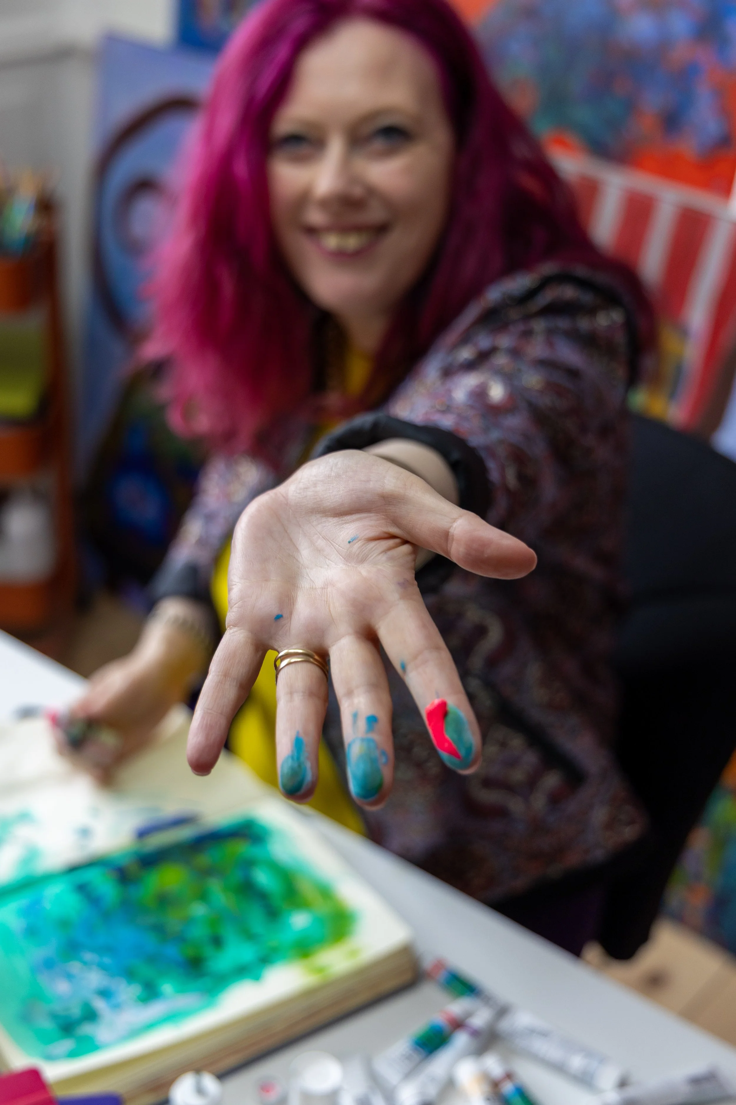 Woman with vibrant pink hair smiling and showing her hand covered in colorful paint, seated at an art table with an open sketchbook and painting supplies around her.