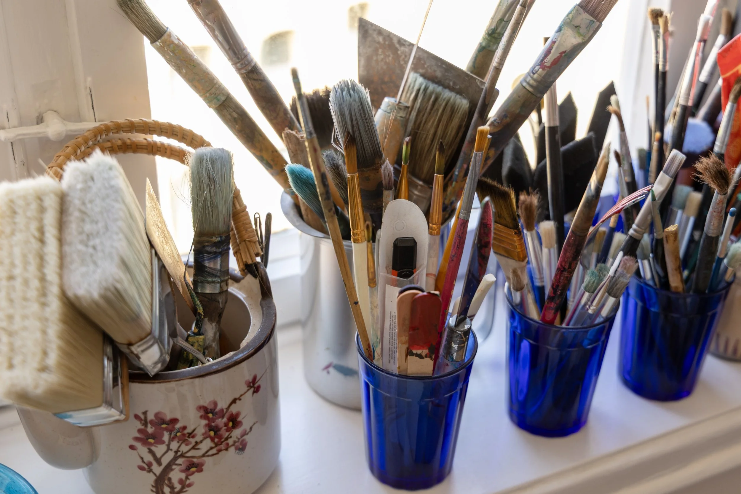 A collection of paintbrushes stored in three blue glass cups and a ceramic mug with a cherry blossom design, placed on a white surface near a window. The brushes vary in size and shape, with some showing signs of use.
