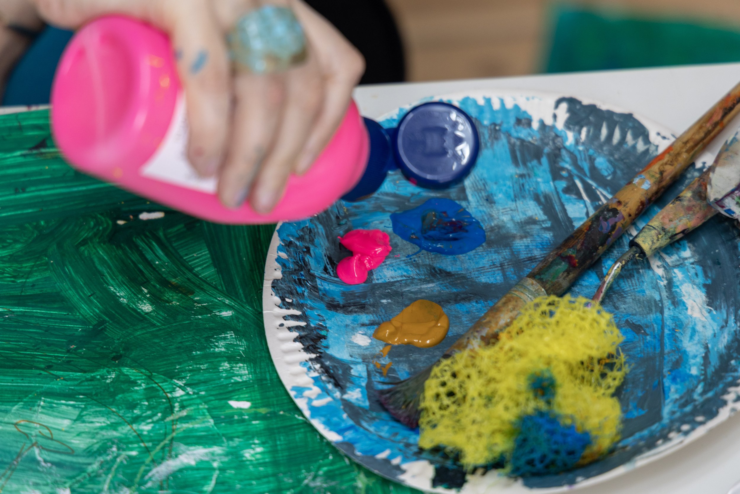 Person pouring pink paint onto a palette of blue, yellow, and pink paint alongside paintbrushes and a painted green surface.