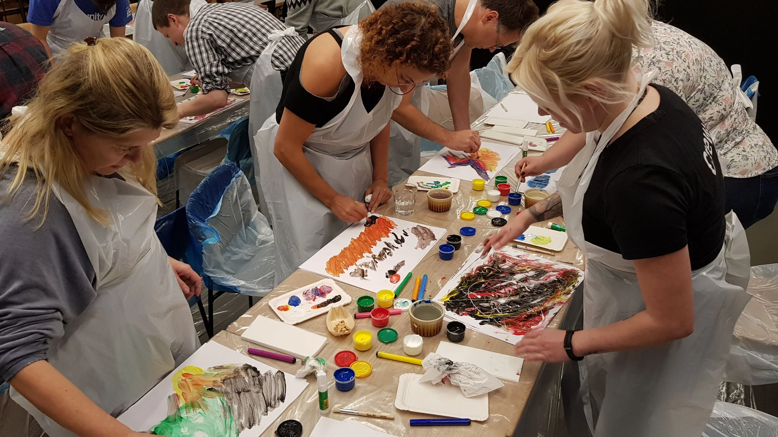 A group of people participating in a painting or art workshop, working on colorful abstract paintings on paper, with various paints, brushes, and art supplies on the table.