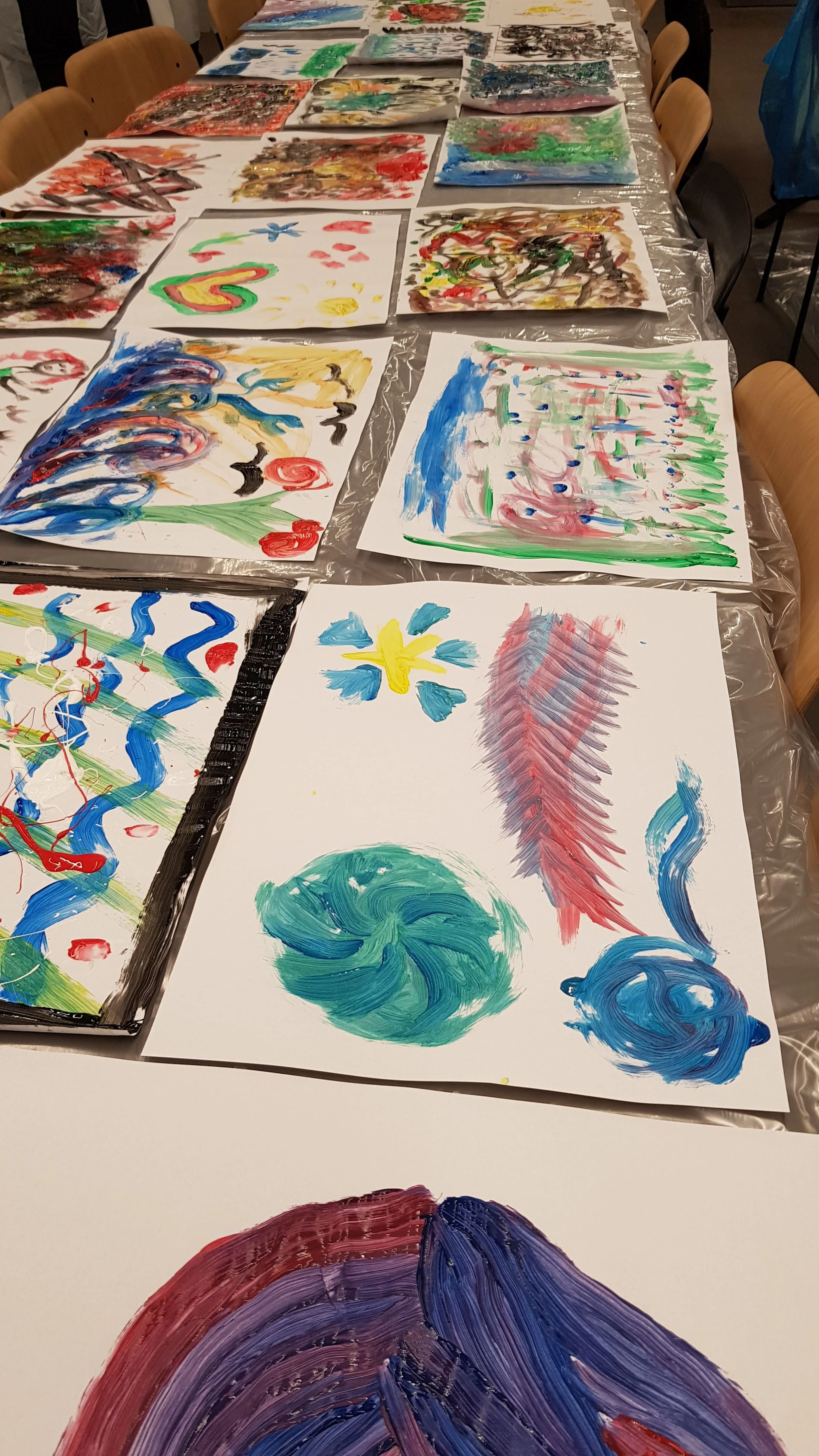Table filled with colorful abstract paintings created with paint and brushes, showcasing vibrant brushstrokes and patterns on large sheets of paper, in an art studio or classroom setting.