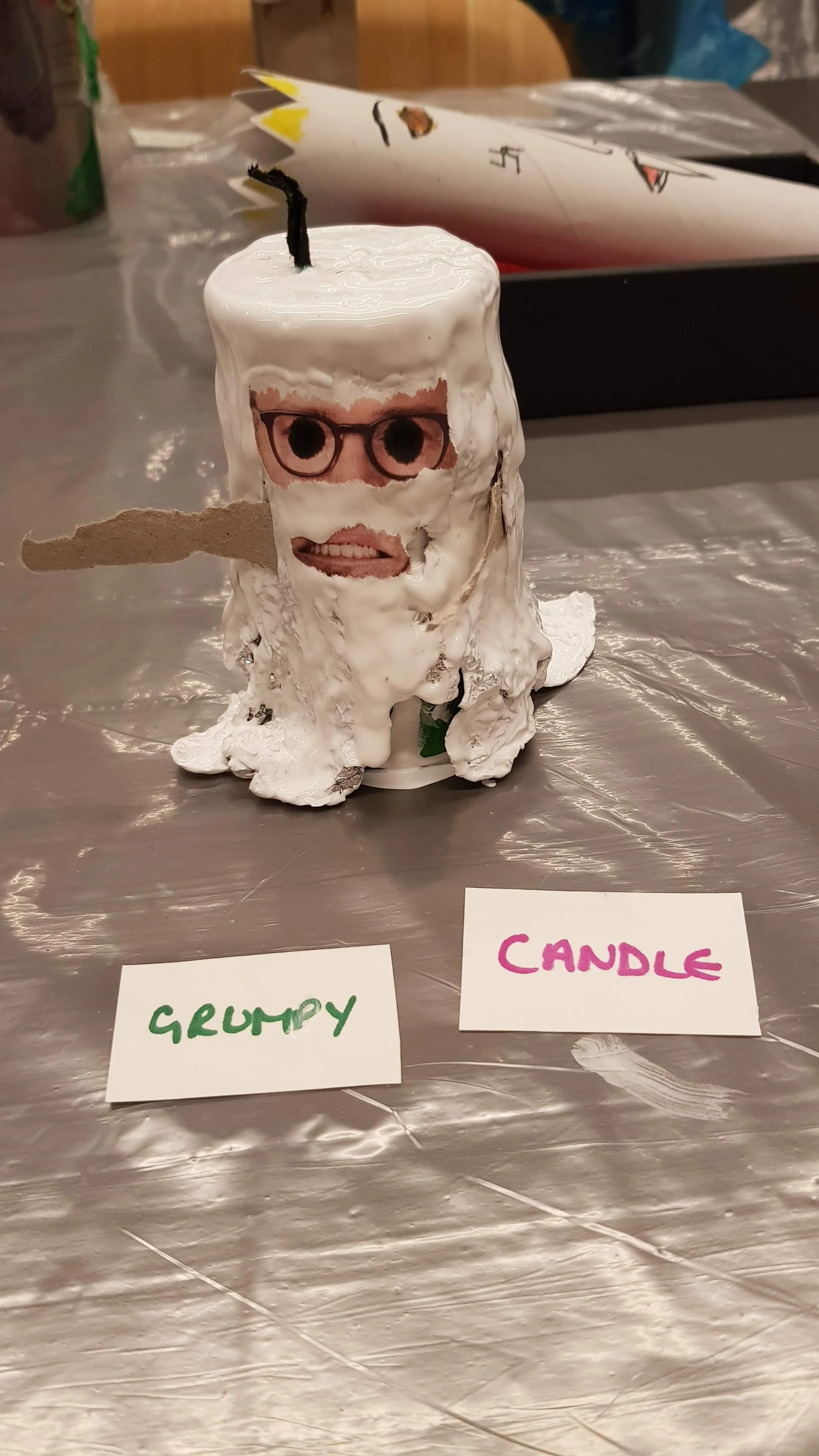 A homemade candle with a human face collage on it, candles wick at the top, and decorated with a paper tongue and glasses. It is labeled 'Grumpy Candle' with a card in front of it.