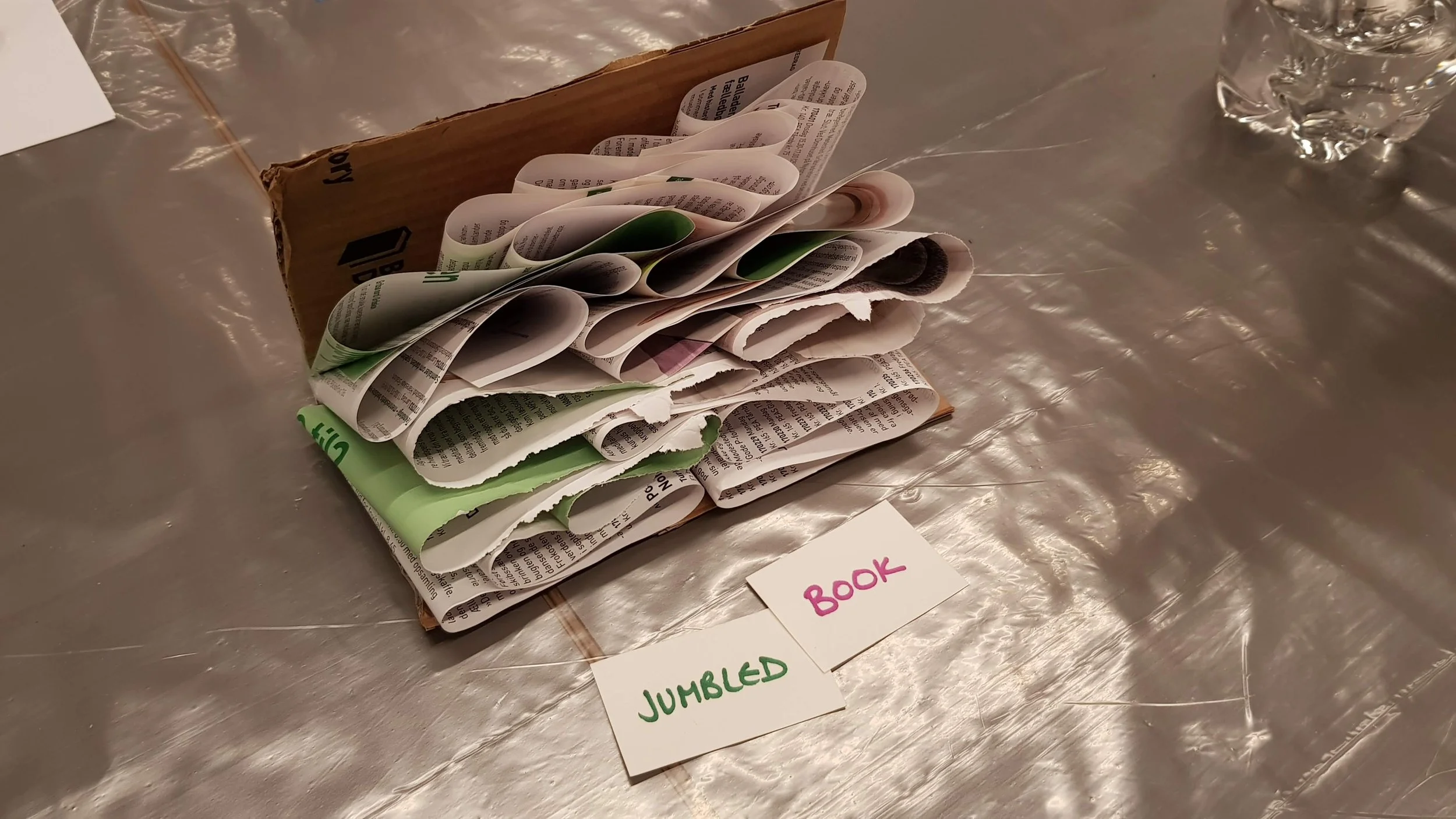 A small cardboard box filled with crumpled and rolled-up pieces of newspapers, placed on a metallic table. Next to the box are two pieces of paper labeled 'JUMBLED' in green and 'BOOK' in pink.