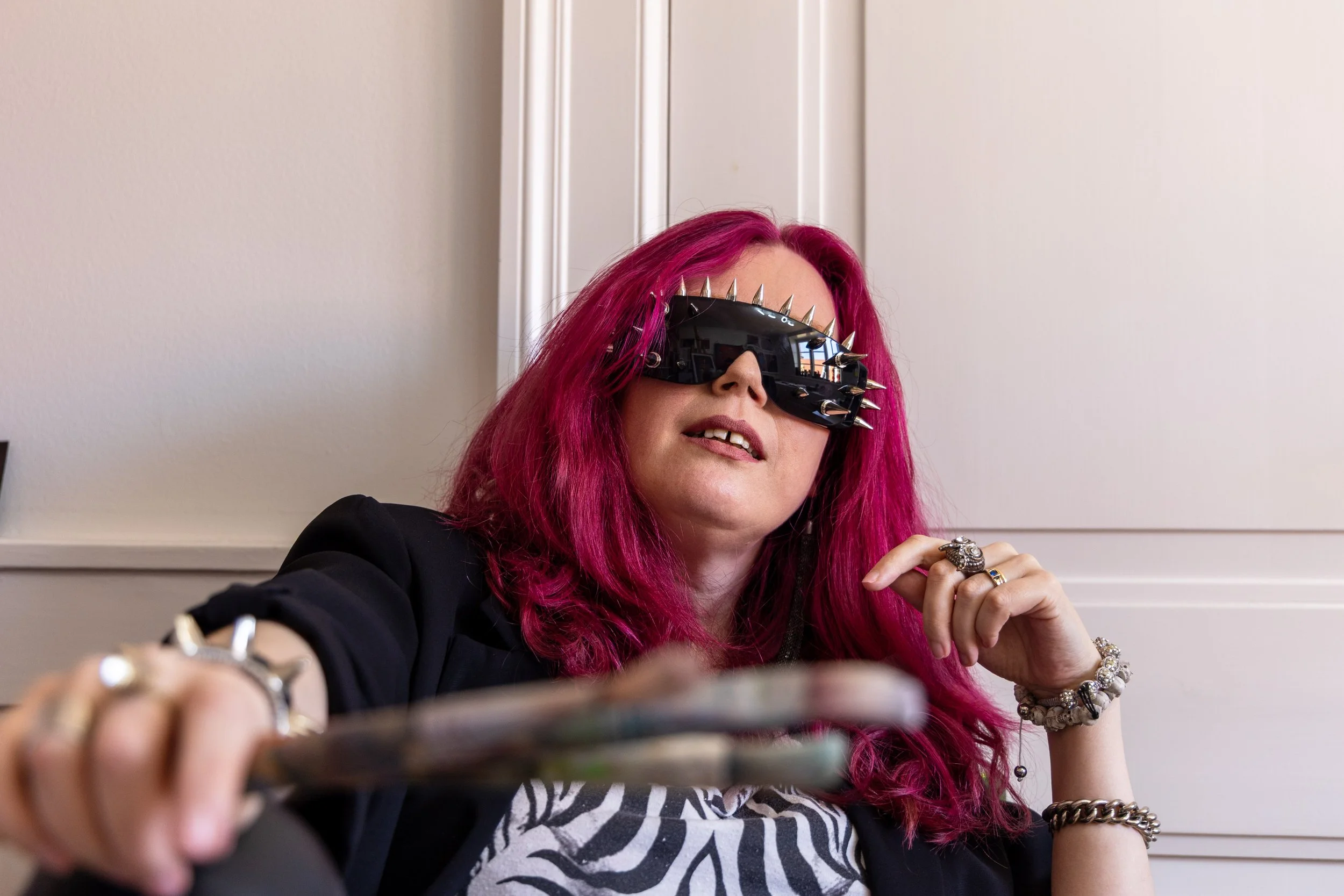 Woman with bright pink hair wearing futuristic sunglasses with spikes, jewelry on her fingers and wrist, sitting indoors against white paneling, taking a selfie with a smartphone.