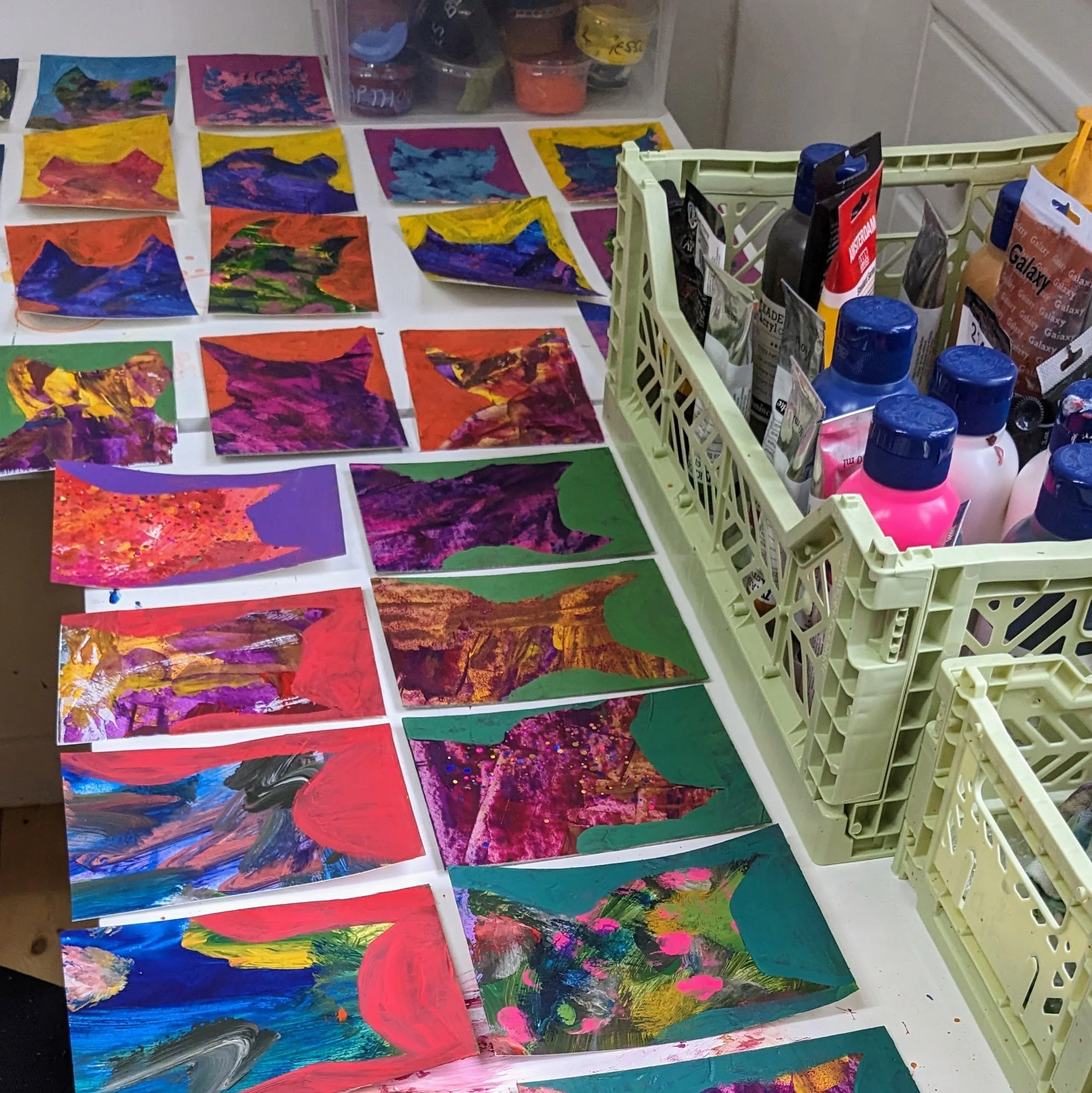 Colorful abstract paintings and artwork laid out on a table with art supplies in a plastic caddy nearby.