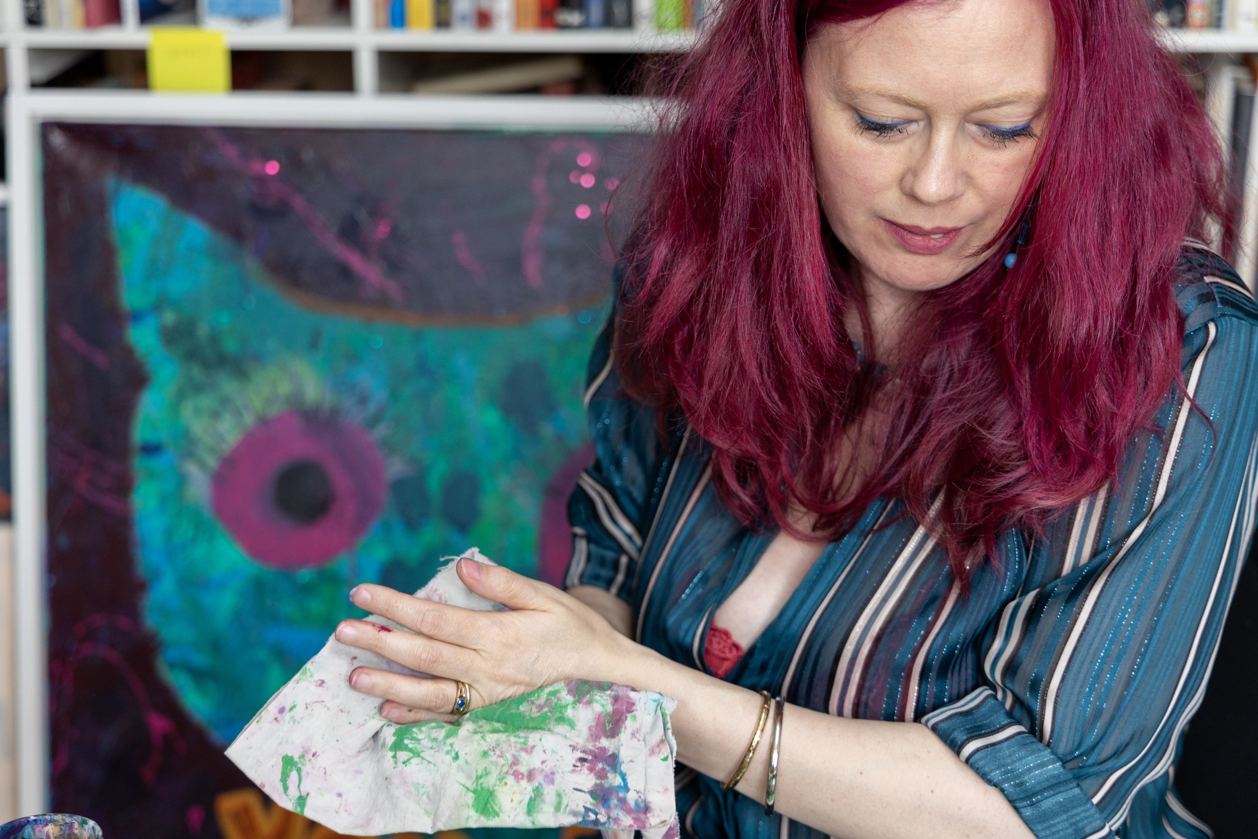 A woman with pink hair creating art, holding a cloth stained with paint, with an abstract colorful painting of a peacock in the background.