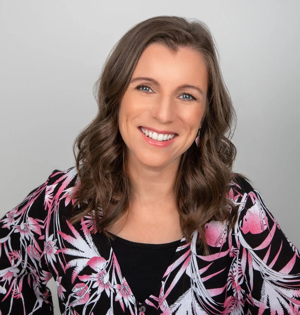 A smiling woman with long wavy brown hair, wearing a black top with pink and white floral and leaf pattern, posing against a plain gray background.