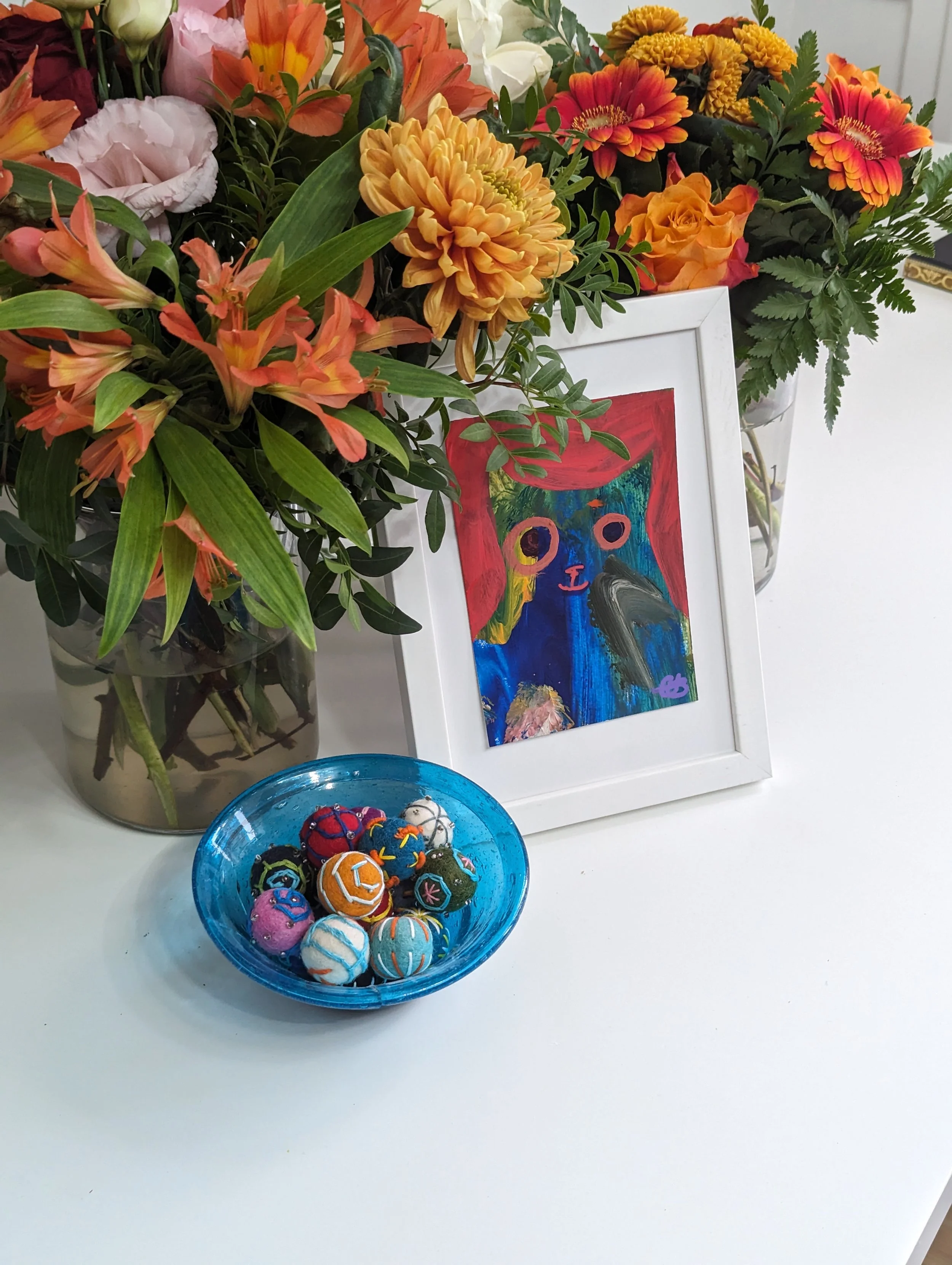 Colorful bouquet of flowers in a vase, framed artwork with abstract face, and a blue bowl with decorative balls on a white surface.