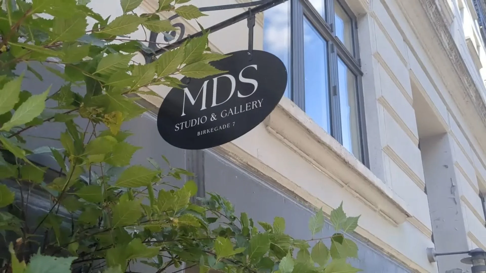 Black oval sign for MDS Studio & Gallery hanging on a building. The sign includes the address Birke Gade 7. Light-colored building with large windows and some green leafy branches in the foreground.