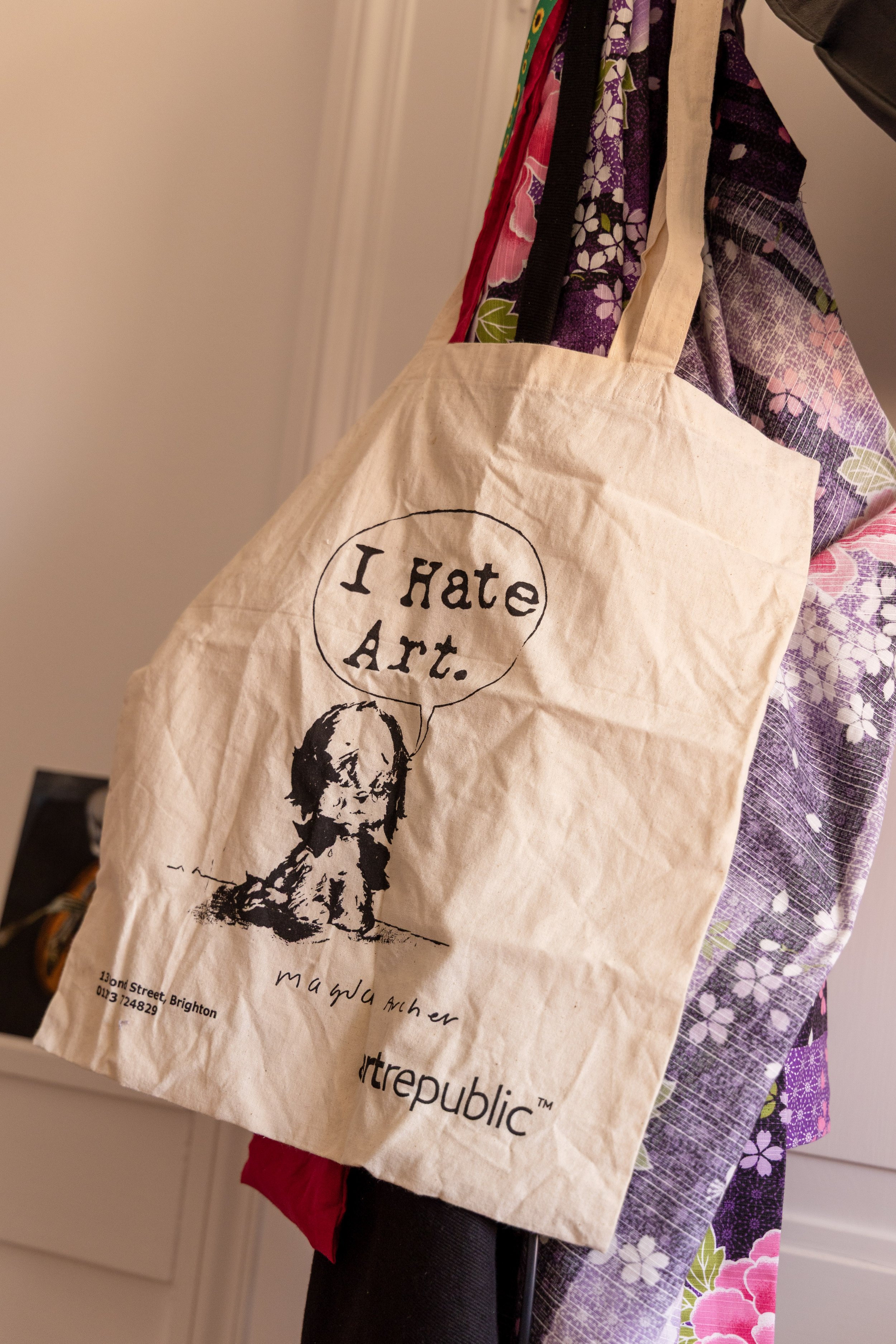 A beige tote bag with a drawing of a girl and the text 'I Hate Art.' The bag is hanging on a coat rack, with colorful fabric or clothing partly visible behind it.