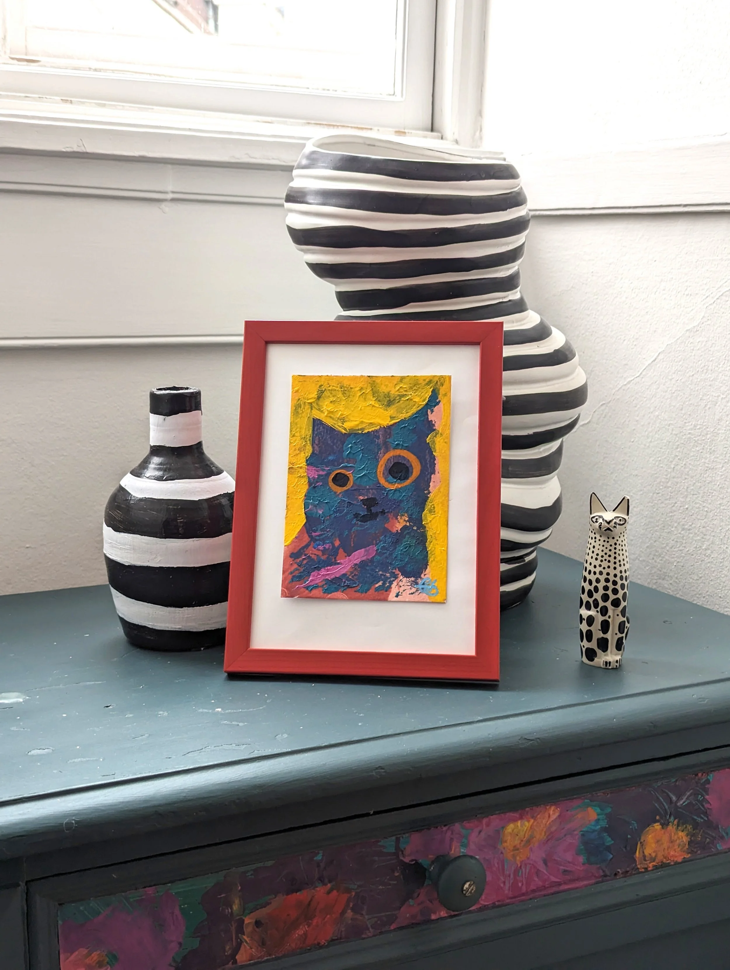 Decorative display on a blue cabinet with striped vases, a colorful cat portrait in a red frame, and a small spotted ceramic cat figurine near a window.