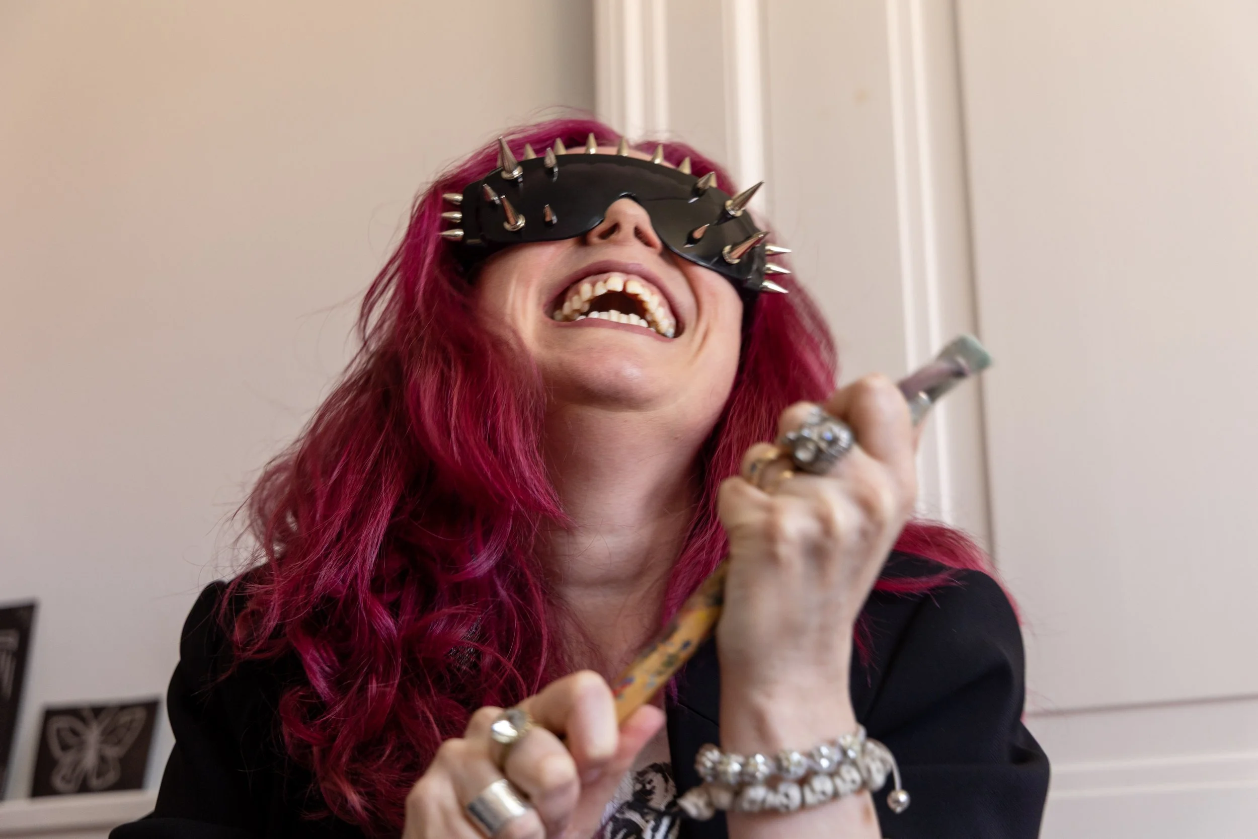 Woman with long pink hair wearing a spiked blindfold, laughing while holding a paintbrush.