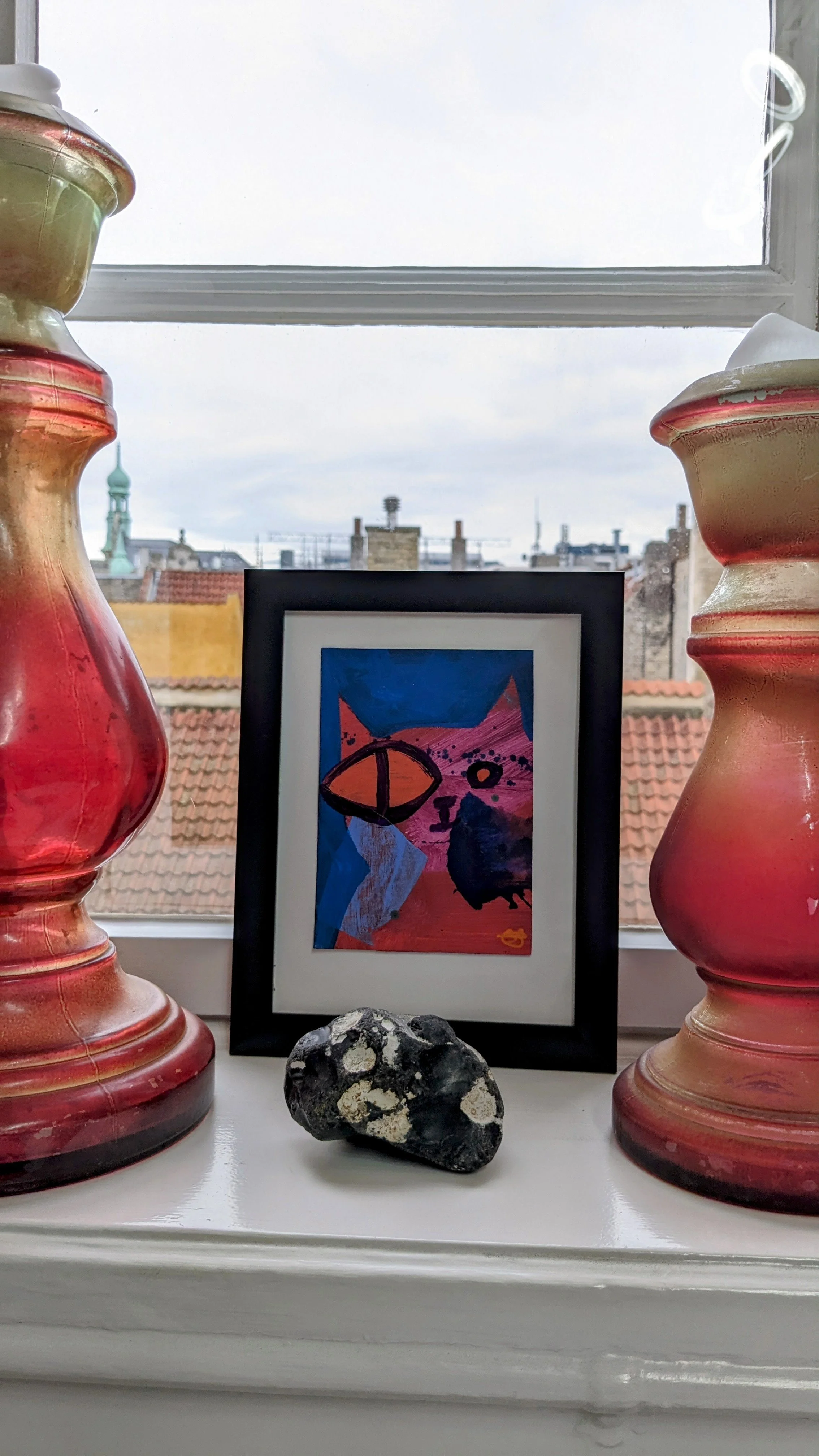 Decorative window sill with two large pink and beige vases, a framed abstract colorful cat artwork, a black rock with white patches, and a view of rooftops and a cloudy sky outside the window.