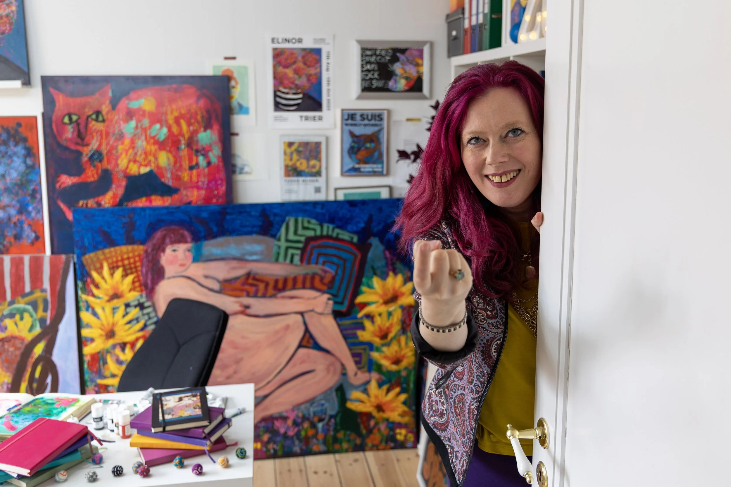 A woman with vibrant pink hair peeks from behind a door, smiling at the camera, in an art studio filled with colorful paintings and art supplies.