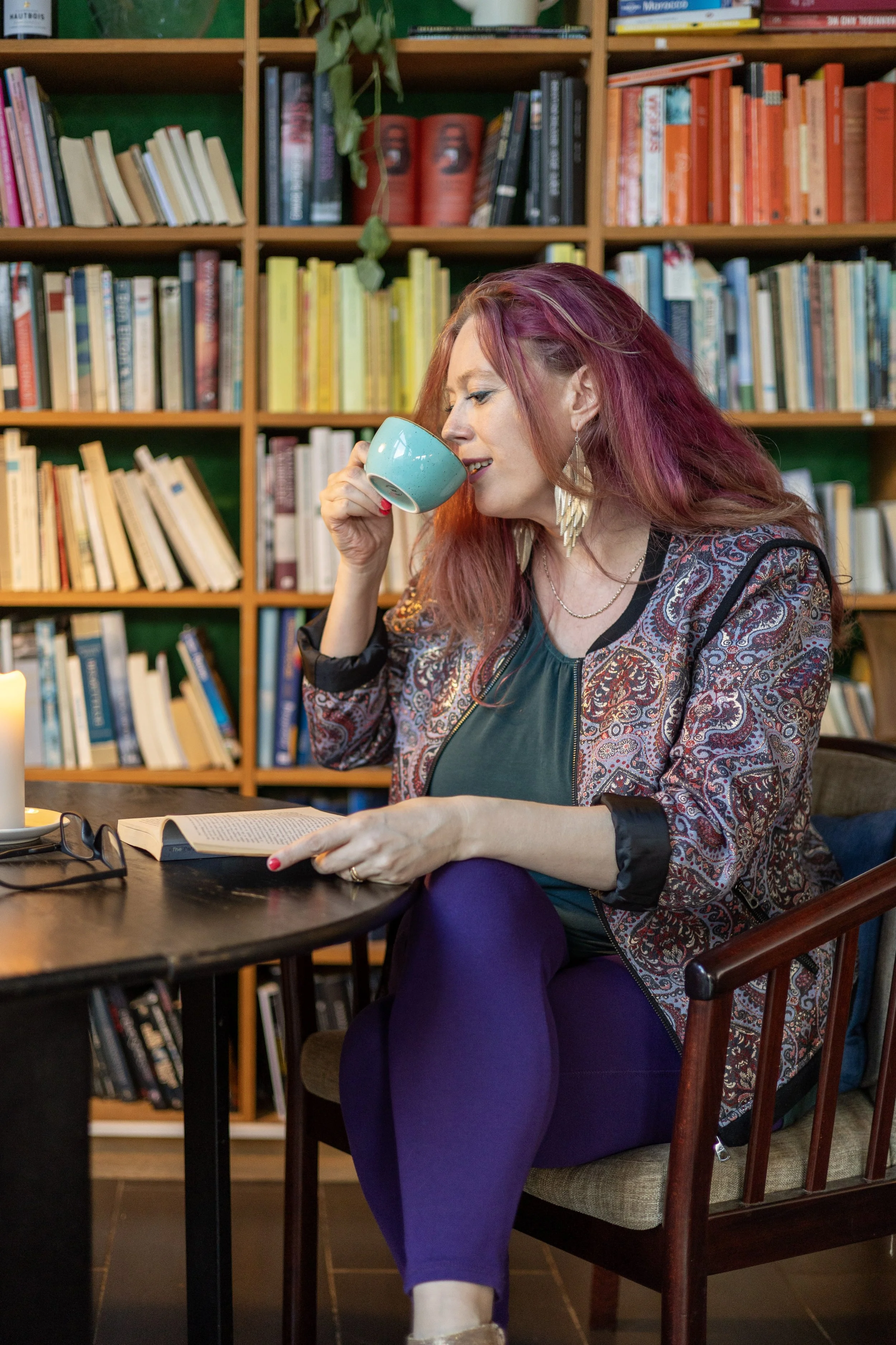A woman with red hair and colorful clothing drinking from a teal cup while sitting at a table with an open book, in front of a bookshelf filled with books.
