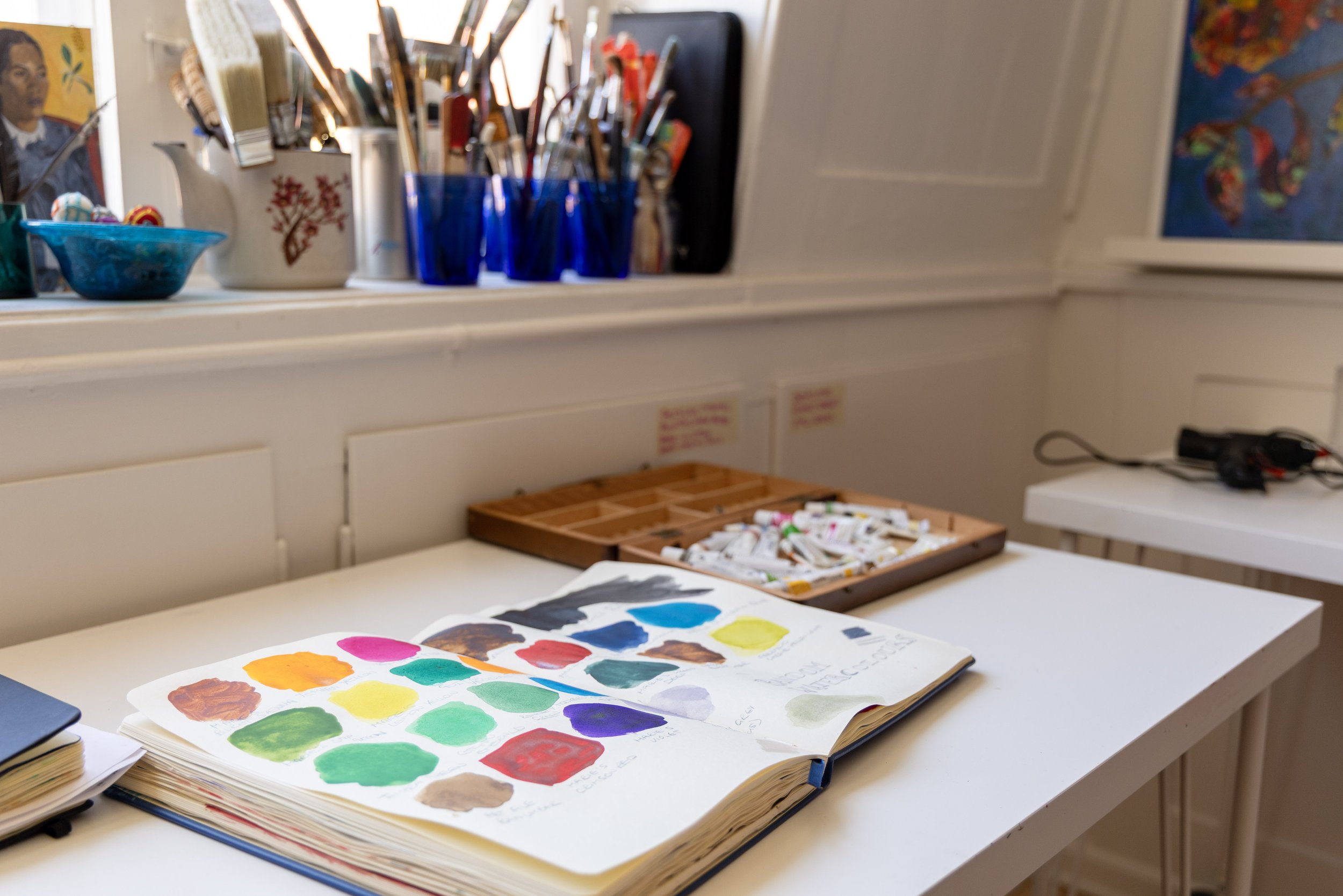Open sketchbook with watercolor paint swatches on a white table in an art studio, with jars of paintbrushes and paints in the background.