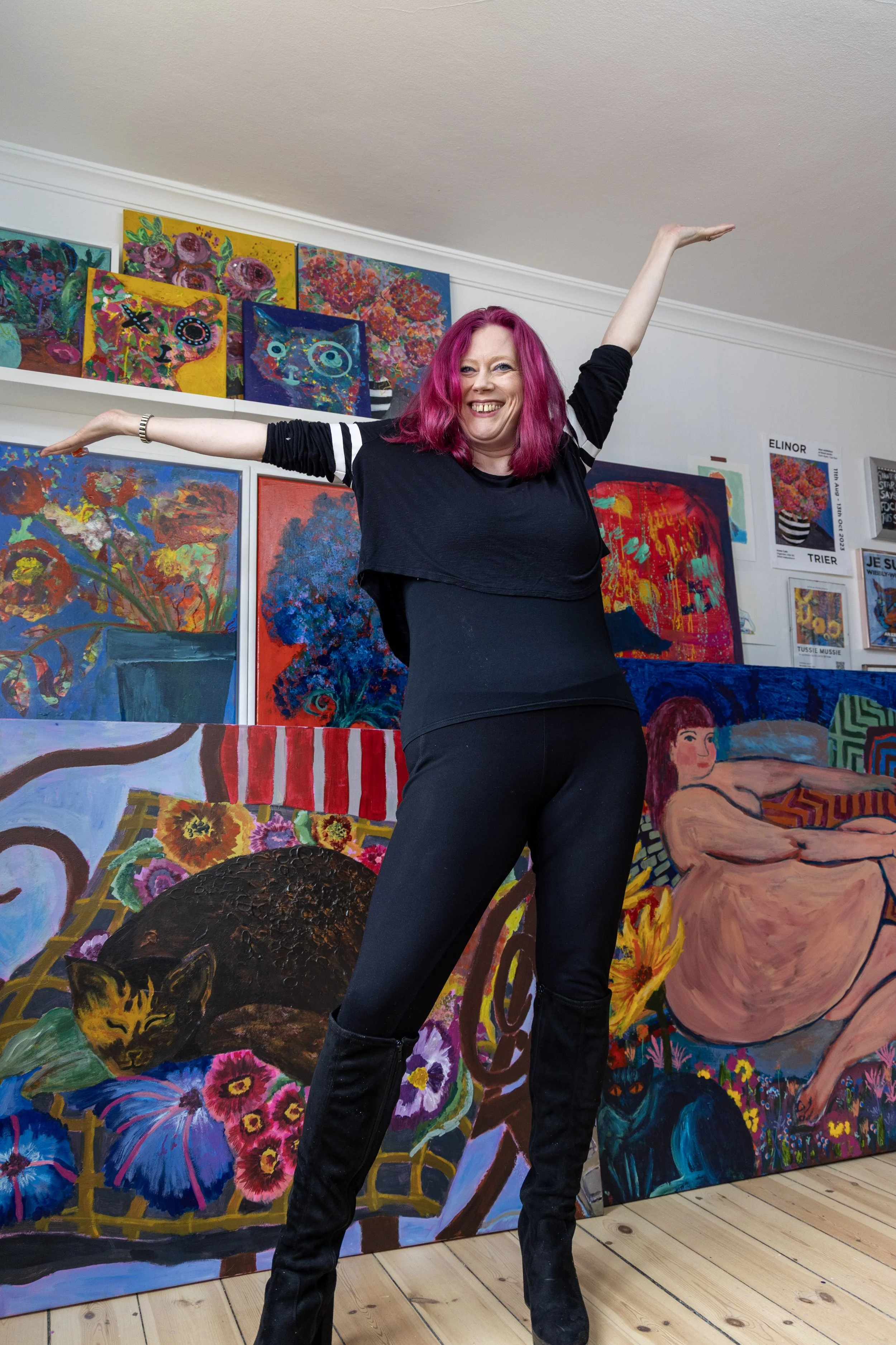 A woman with pink hair smiling and stretching her arms in an art gallery with colorful paintings of flowers, cats, and a nude woman in the background.