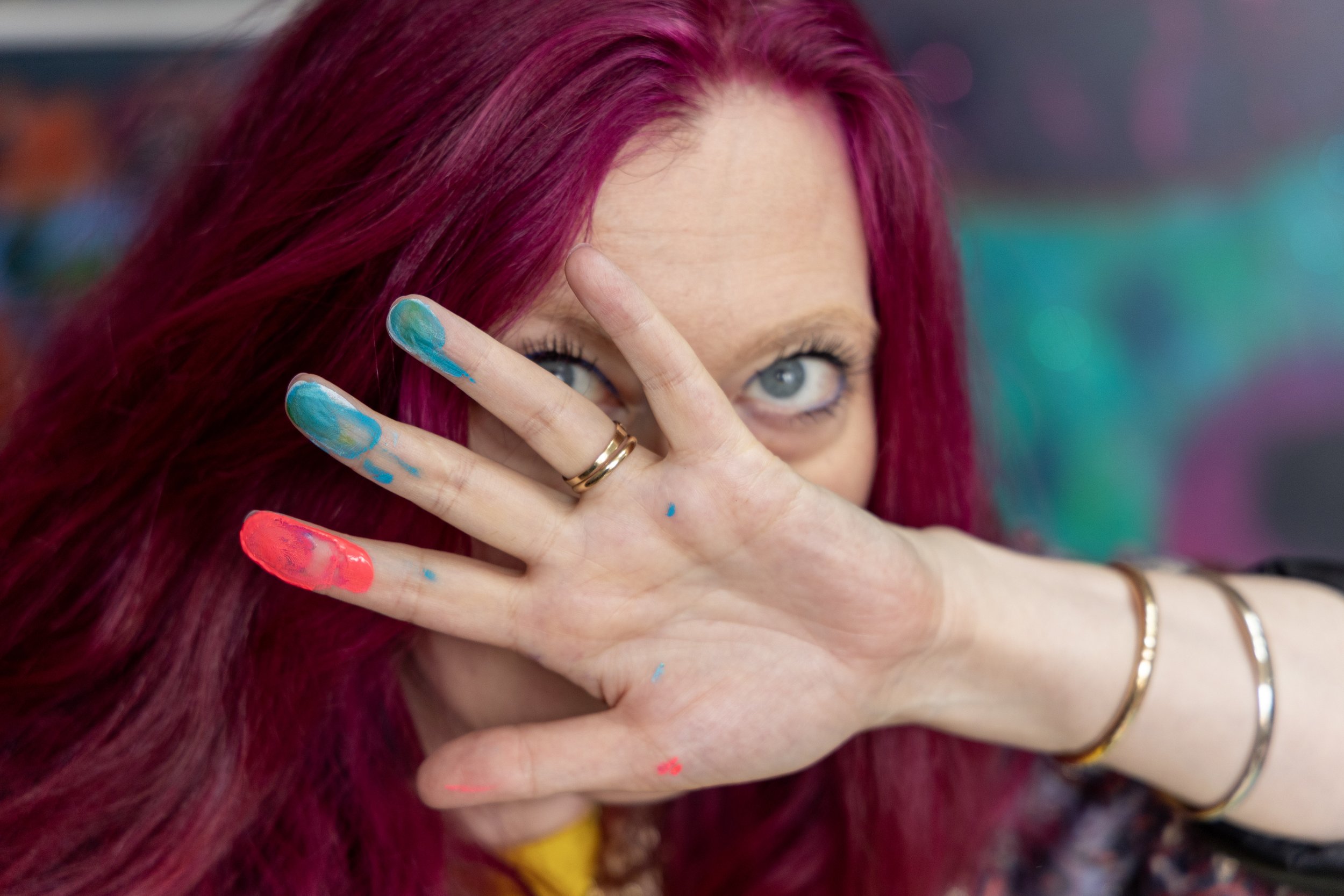 A woman with bright pink hair covering part of her face with her hand, which has colorful paint smudges on the fingers, and her piercing blue eyes visible behind the hand.