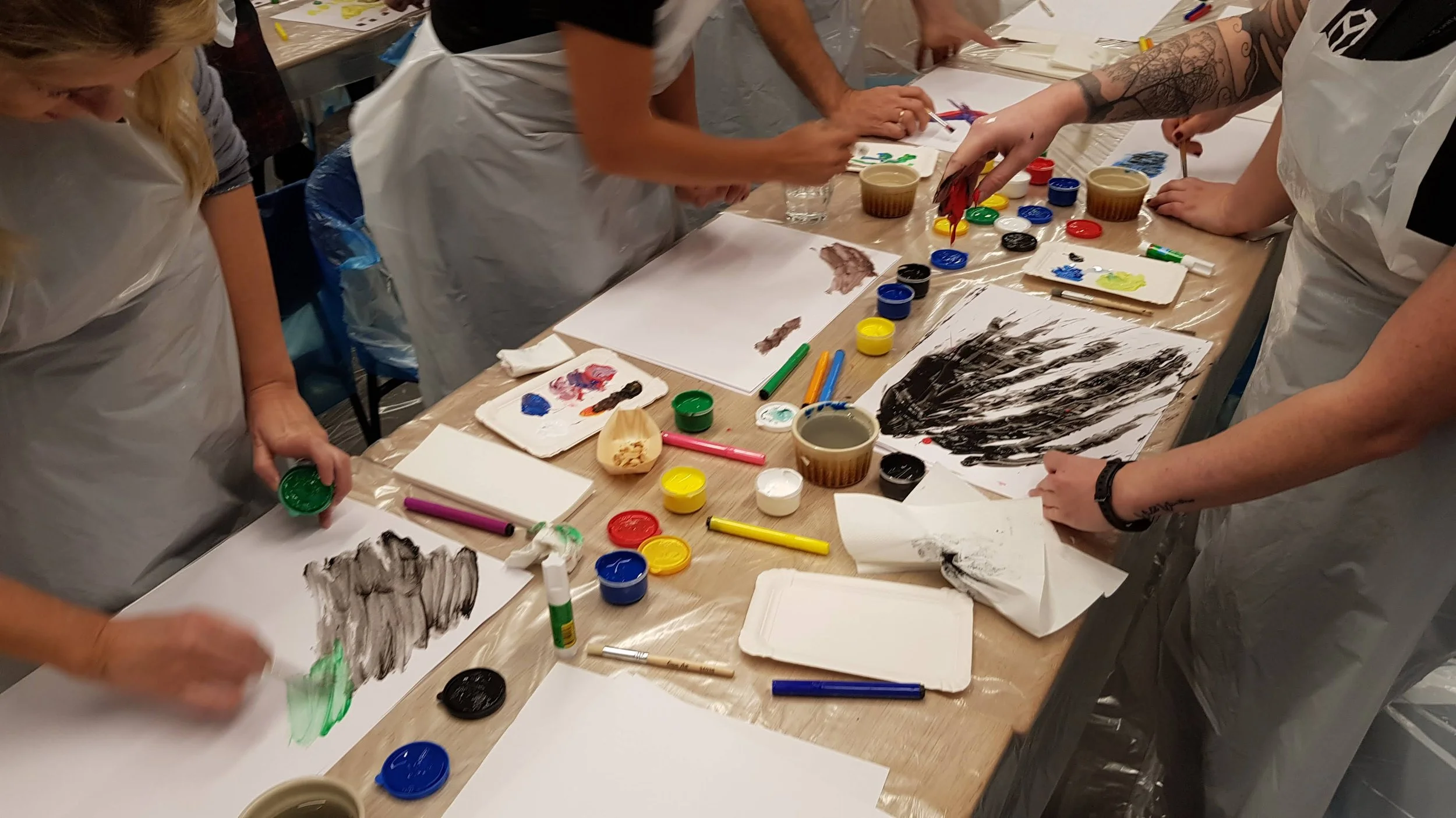 People engaged in a painting activity at a table covered with art supplies like paints, brushes, and paper, wearing white aprons.