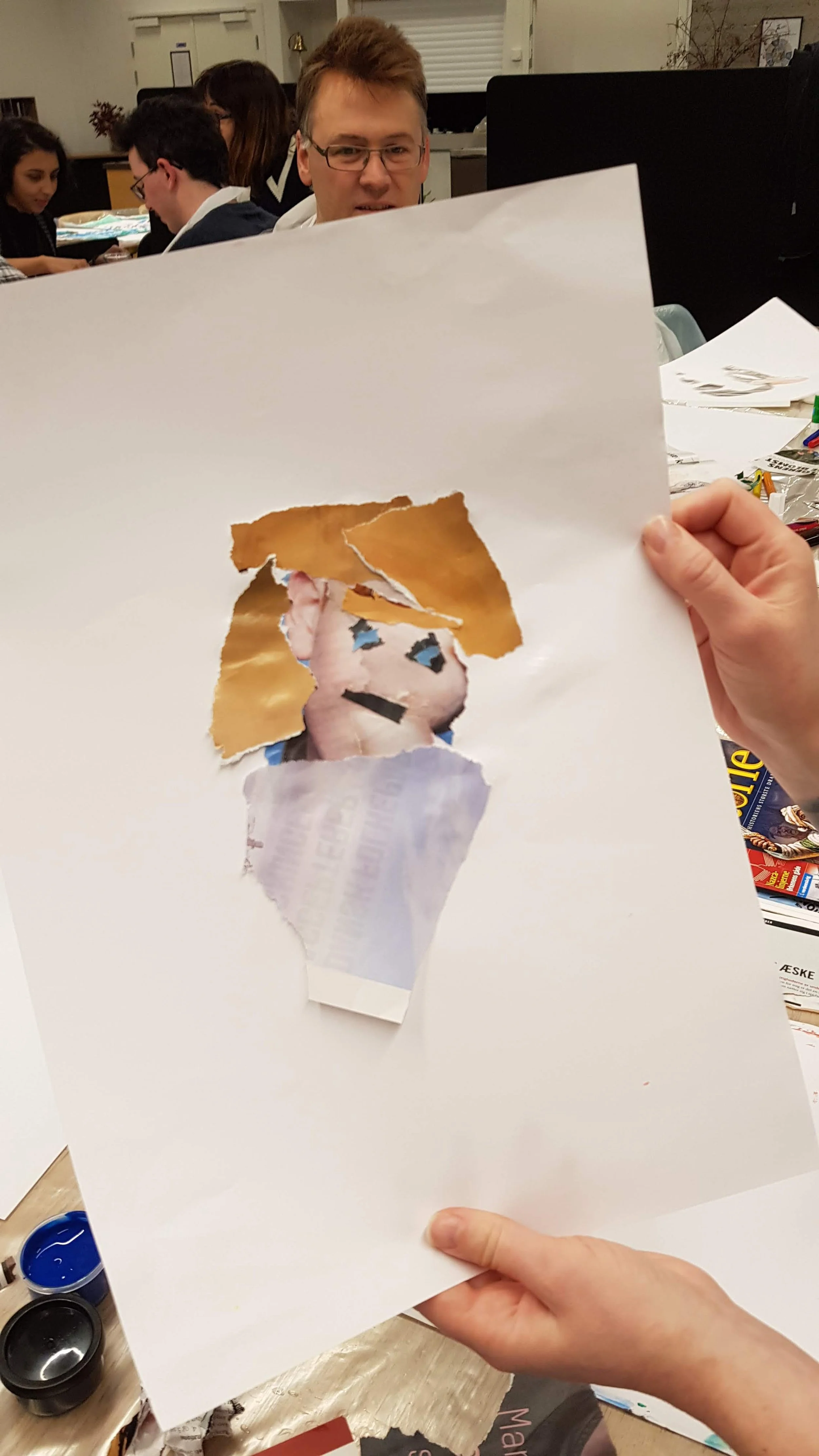 A person holds a torn piece of paper with a partial image of a cartoon character's face, featuring blue eyes, black eyebrows, and a light-colored complexion. In the background, several people are seated at a table in a room with white walls and a win