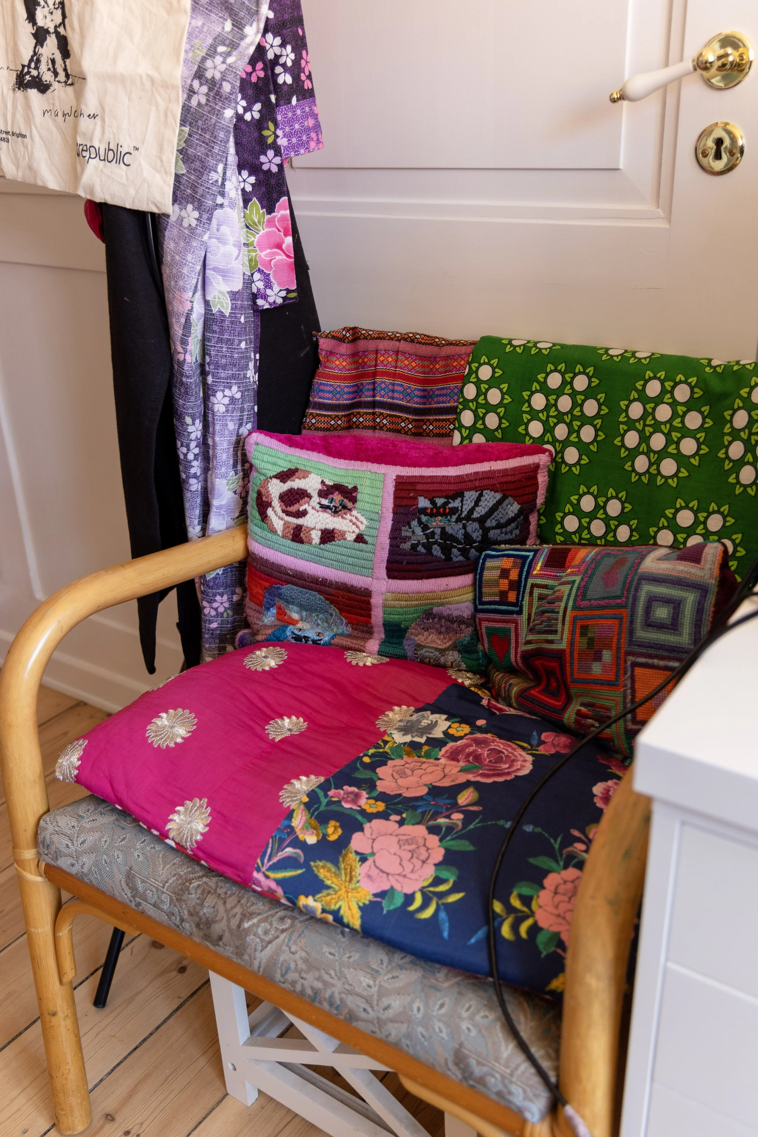A cozy wooden bench with floral and patterned pillows, a pink floral cushion, and a fabric cover, set against a white wall with a white cabinet nearby. Clothes hang on a hook or rack beside the bench.
