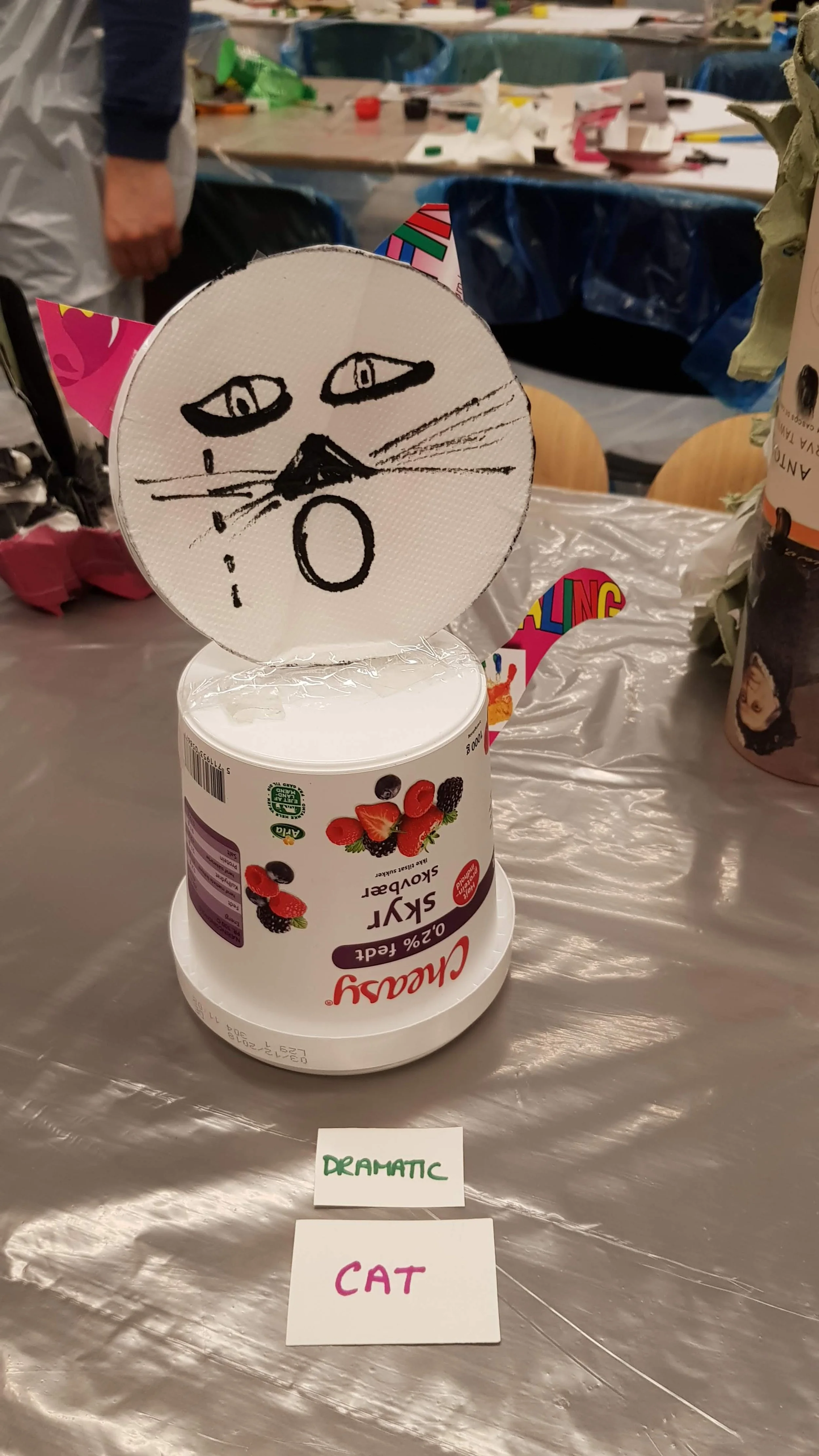 A yogurt container with a hand-drawn cat face and ears on a paper plate, placed on a table covered with plastic. Two small signs in front say 'Dramatic' and 'Cat'.