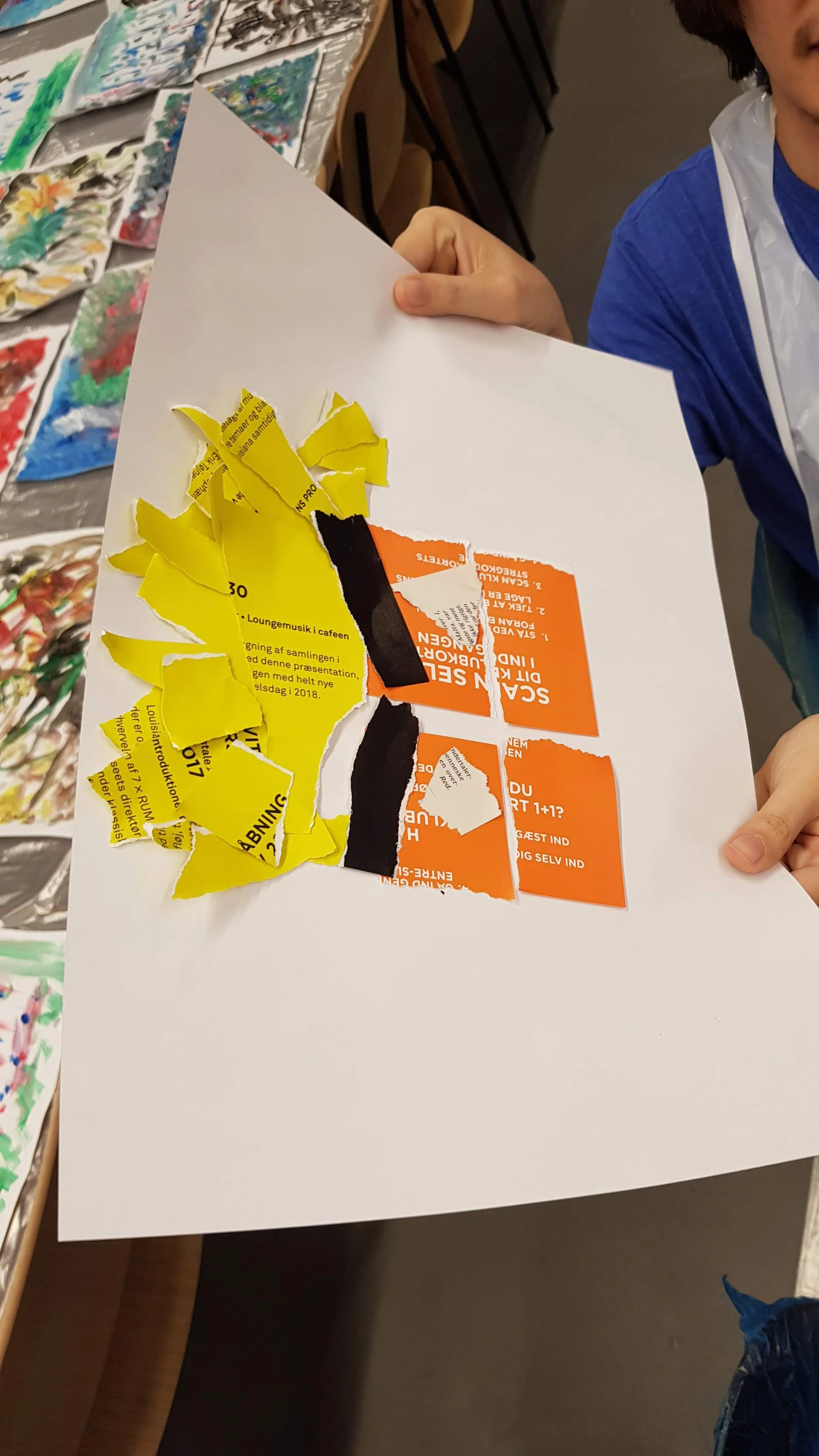 Hand holding a white sheet of paper with a collage of torn yellow, black, white, and orange paper pieces, with an art table and colorful paintings in the background.