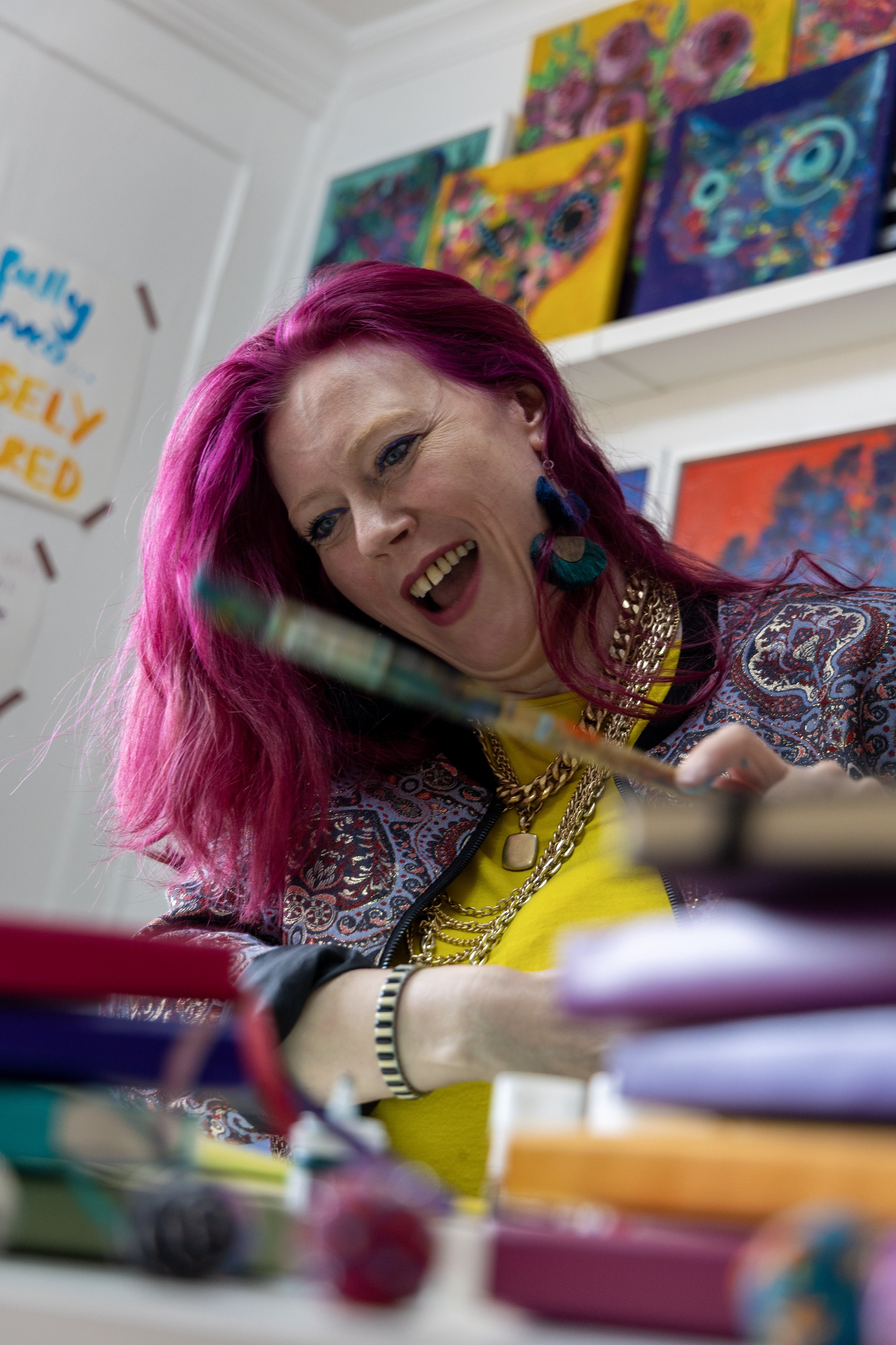 A woman with vibrant pink hair, wearing earrings, layered necklaces, and a colorful jacket, is smiling and engaging with her workspace that has stacks of colorful notebooks or papers, with abstract paintings hanging on the wall behind her.