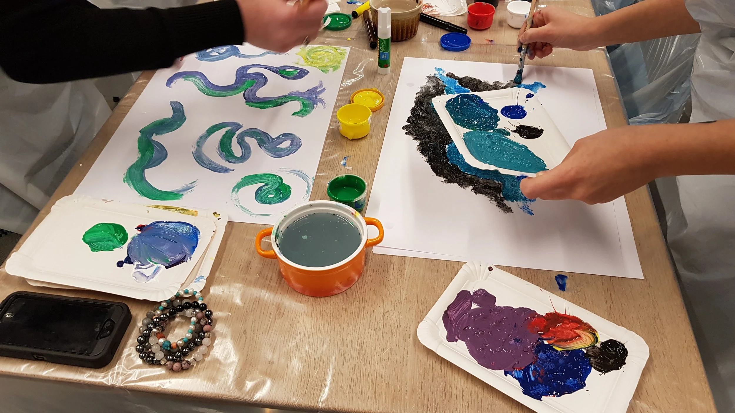 People painting on paper with various colors, using brushes and palettes, surrounded by cups of water and art supplies on a wooden table.