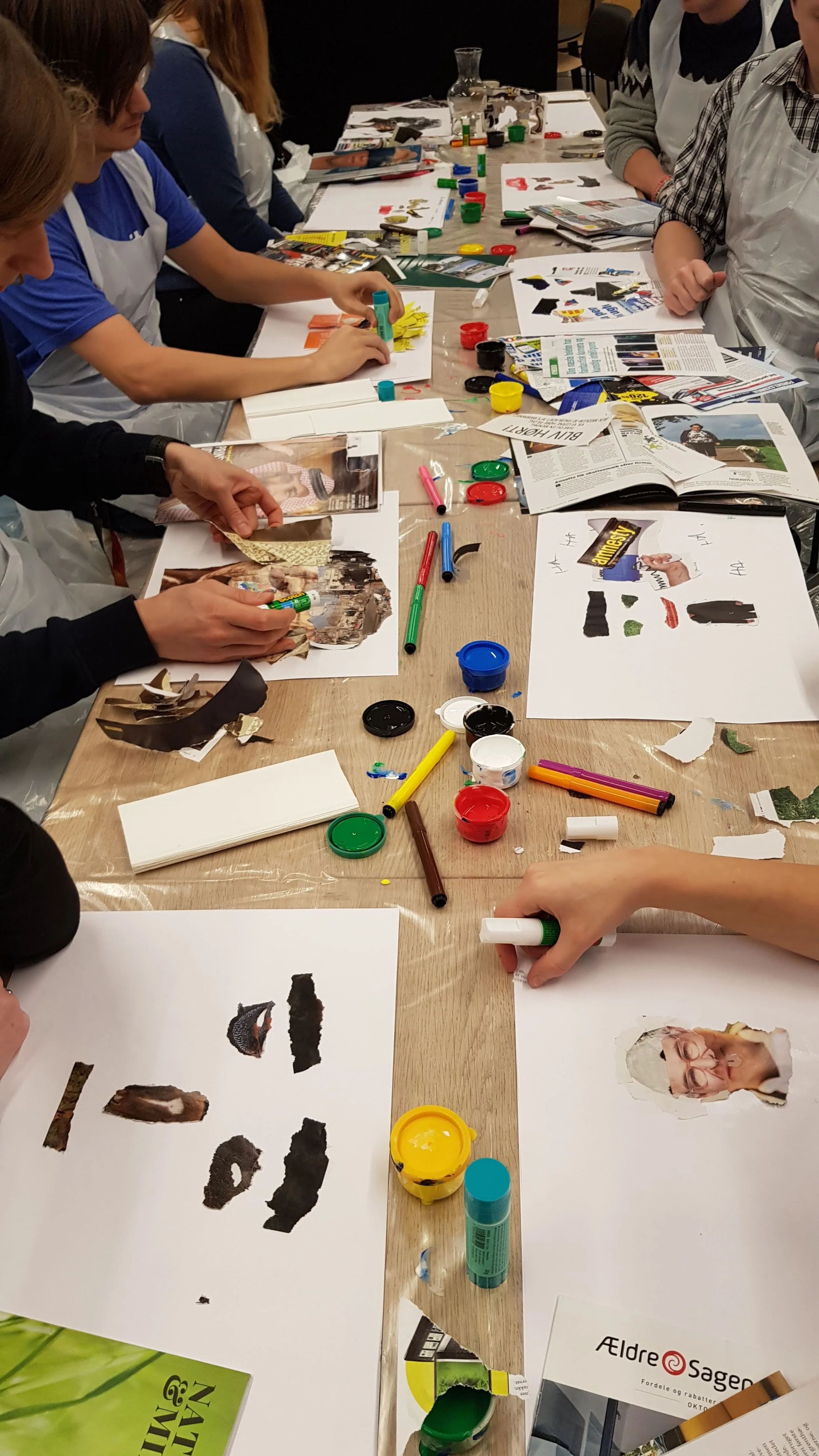 Group of people sitting around a table engaged in collage art making, with magazines, paper, glue, markers, and paint on the table.