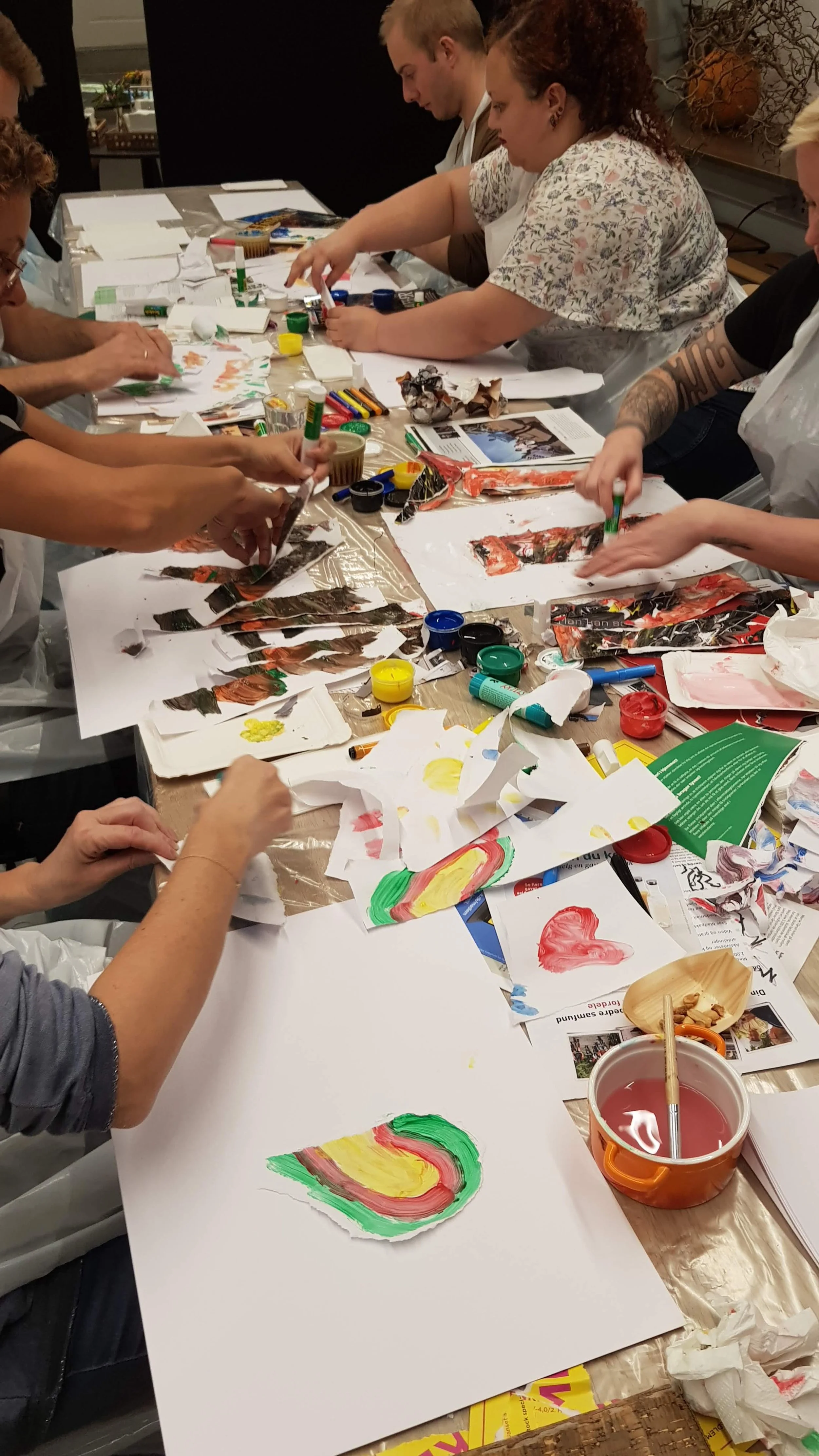 People gathered around a table creating colorful artwork with paint, paper, and markers in a group art activity.